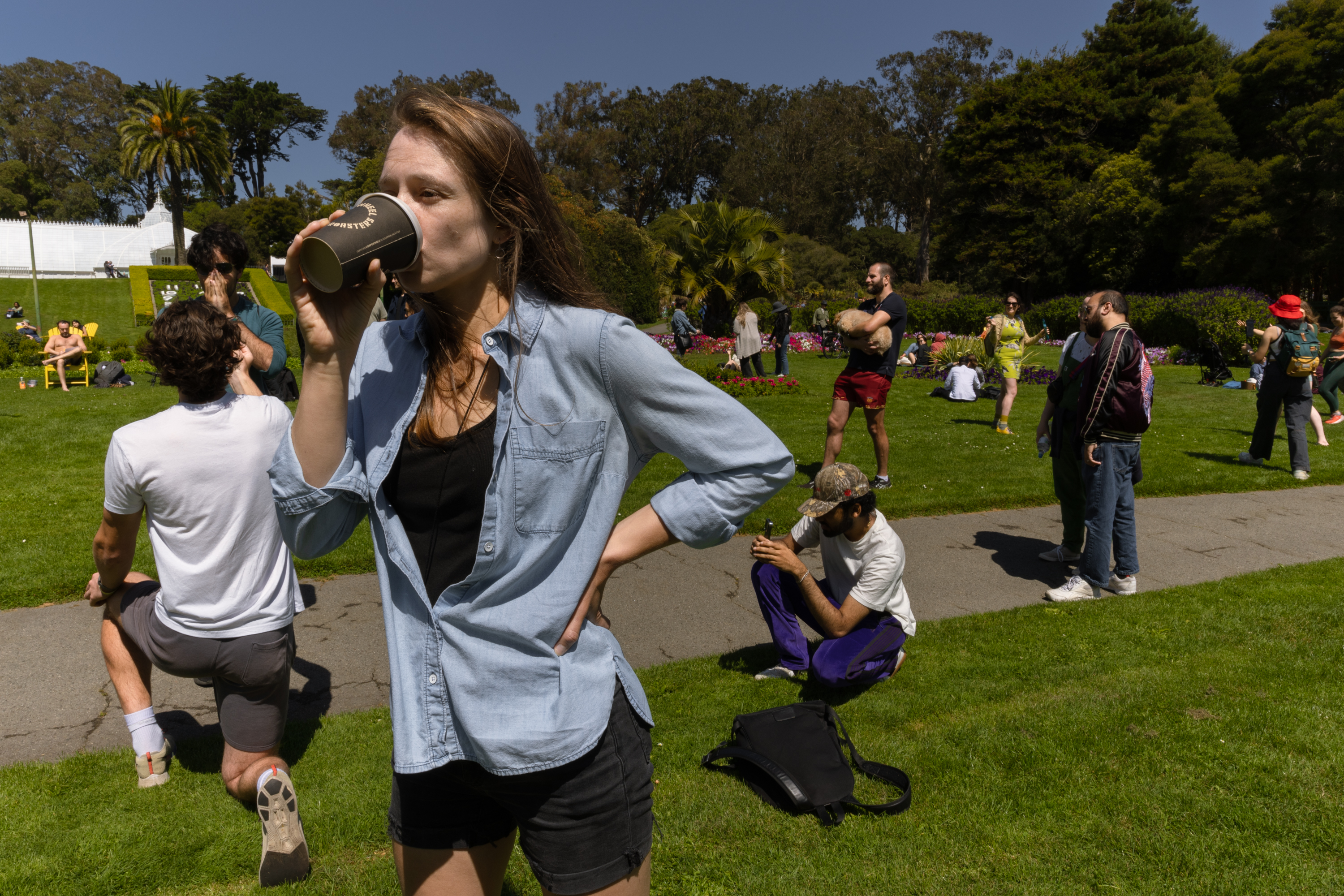 A woman wearing a denim shirt drinks from a cup in a vibrant park, while various people around engage in different activities such as talking, photographing, and lounging.