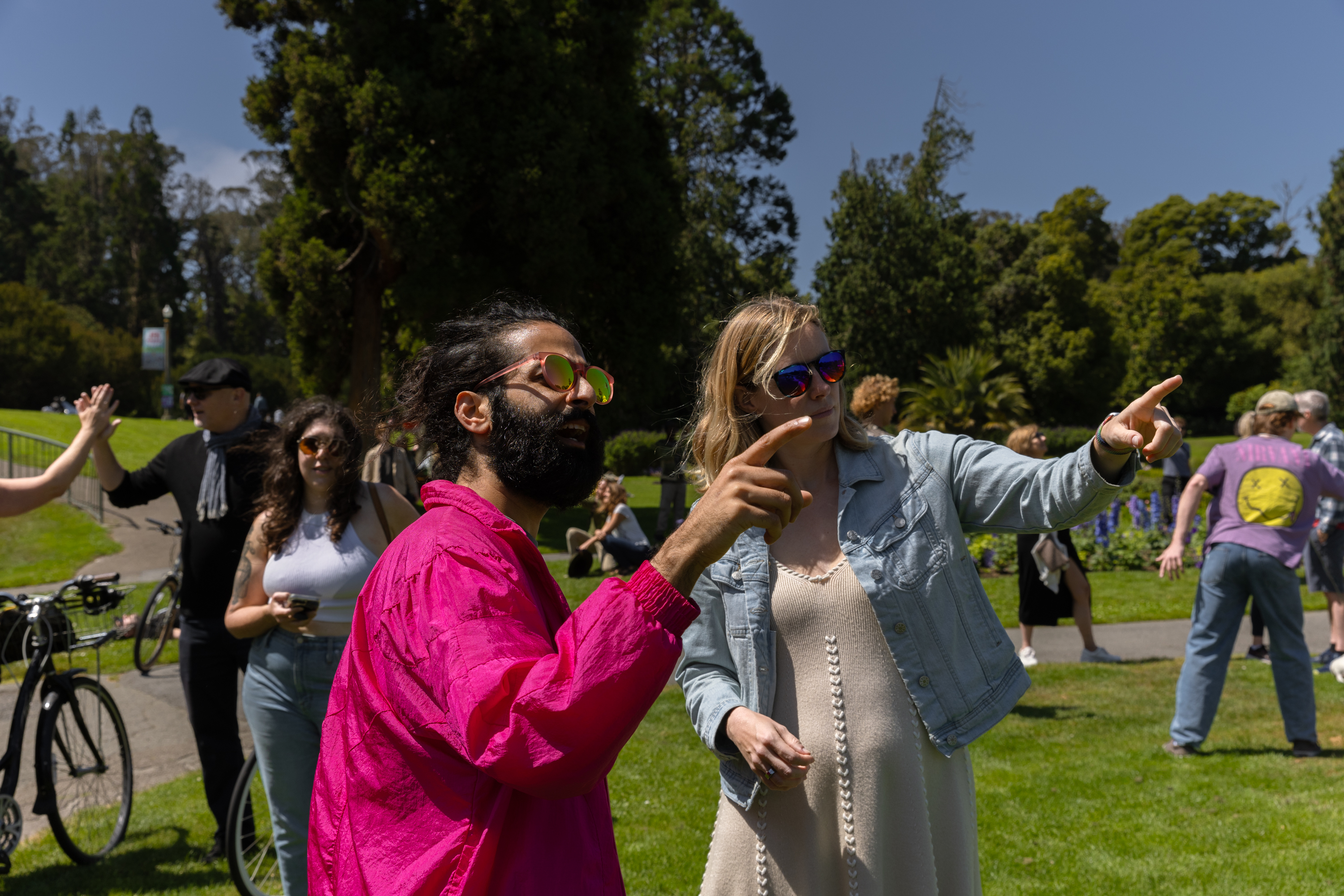 Two people in sunglasses are pointing at something in a park. Others are in the background, with trees and a blue sky visible.