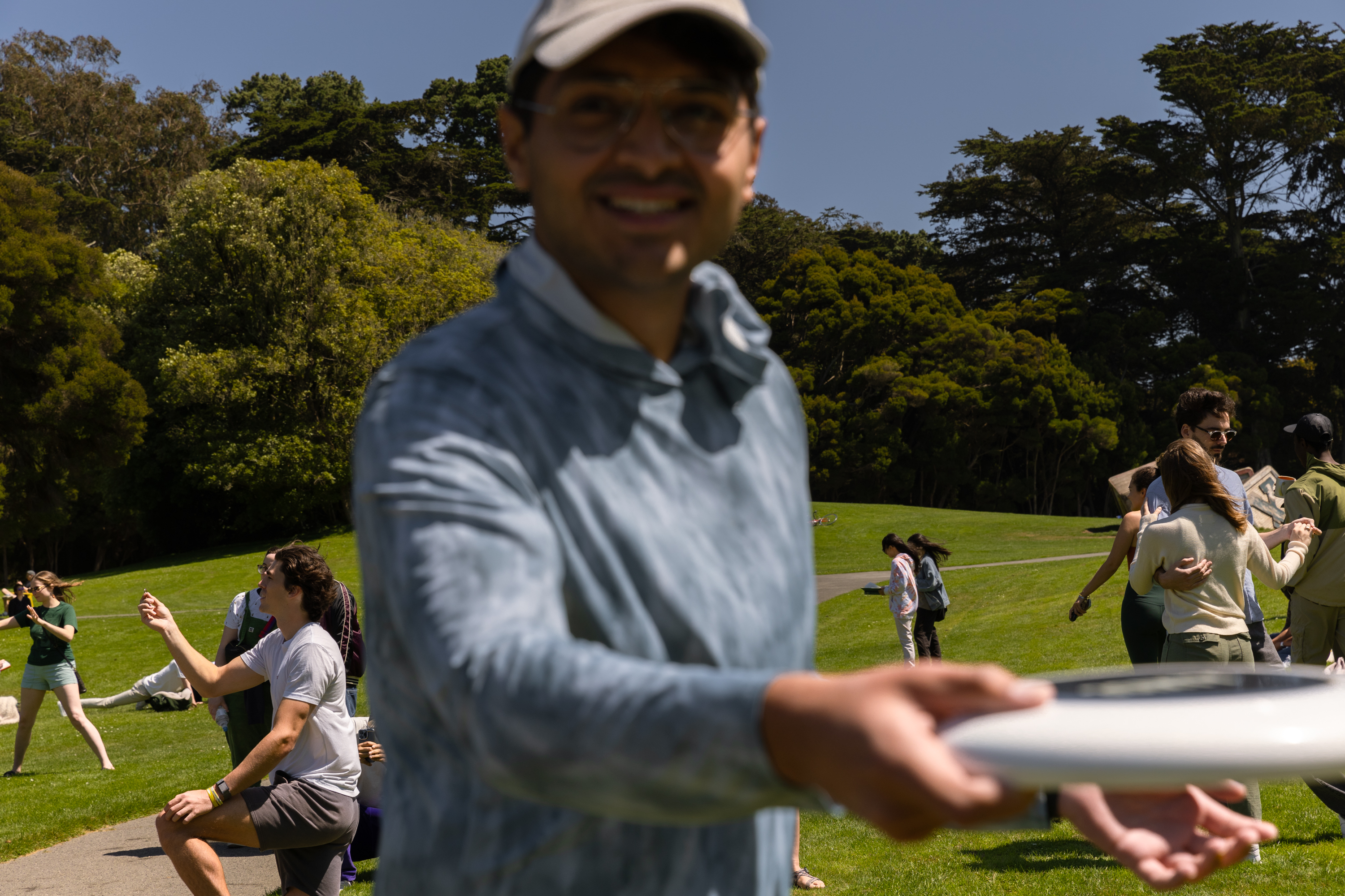 In a sunlit park, a man wearing a cap and glasses smiles, holding a frisbee close to the camera. Behind him, people enjoy various activities on the grassy field.