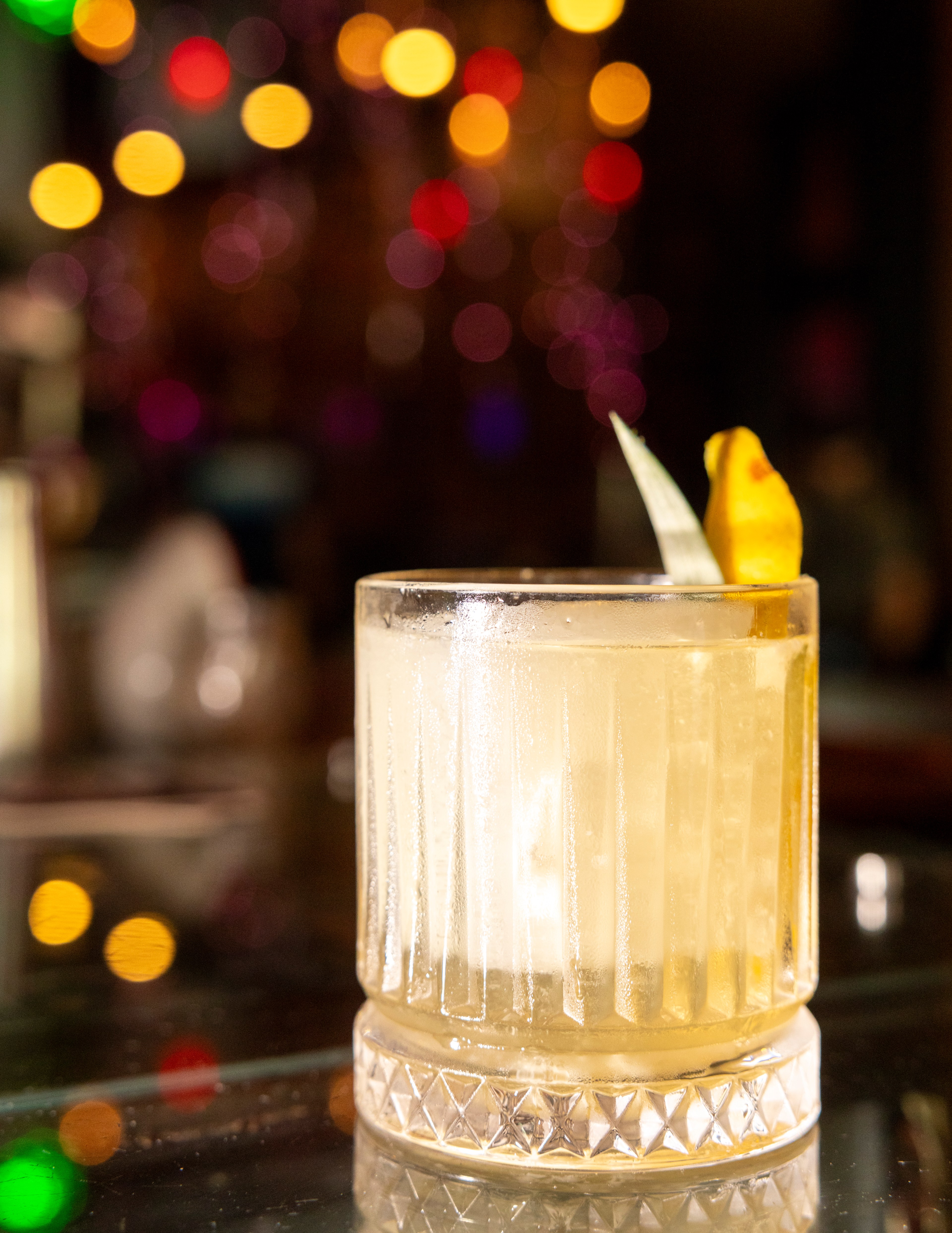 A frosty cocktail in a textured glass with a lemon twist garnish sits on a reflective surface, with colorful out-of-focus lights in the background.