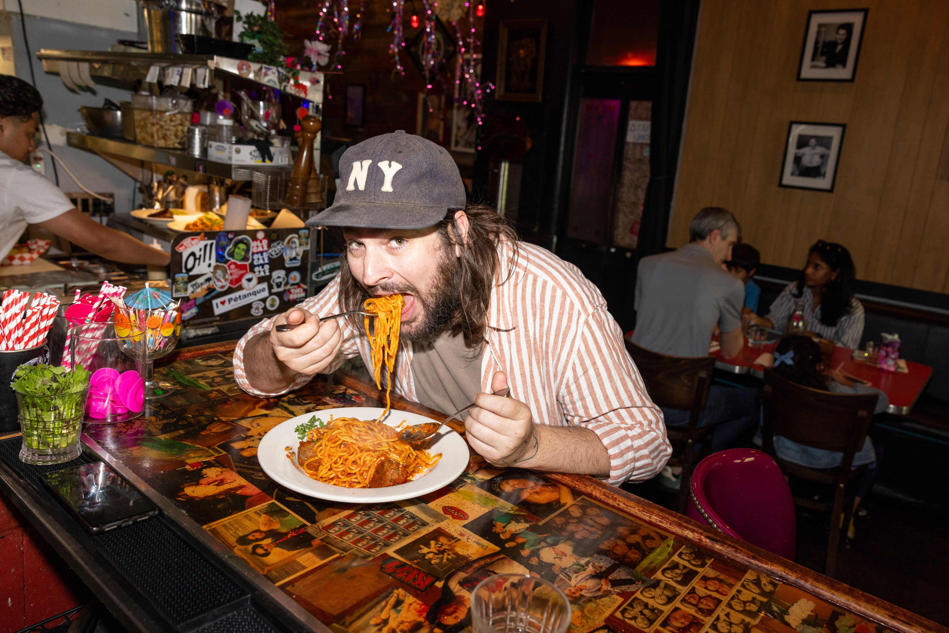 A man wearing a cap labeled "NY" is enthusiastically eating spaghetti at a colorful restaurant counter, with people seated in the background and a busy kitchen seen behind him.