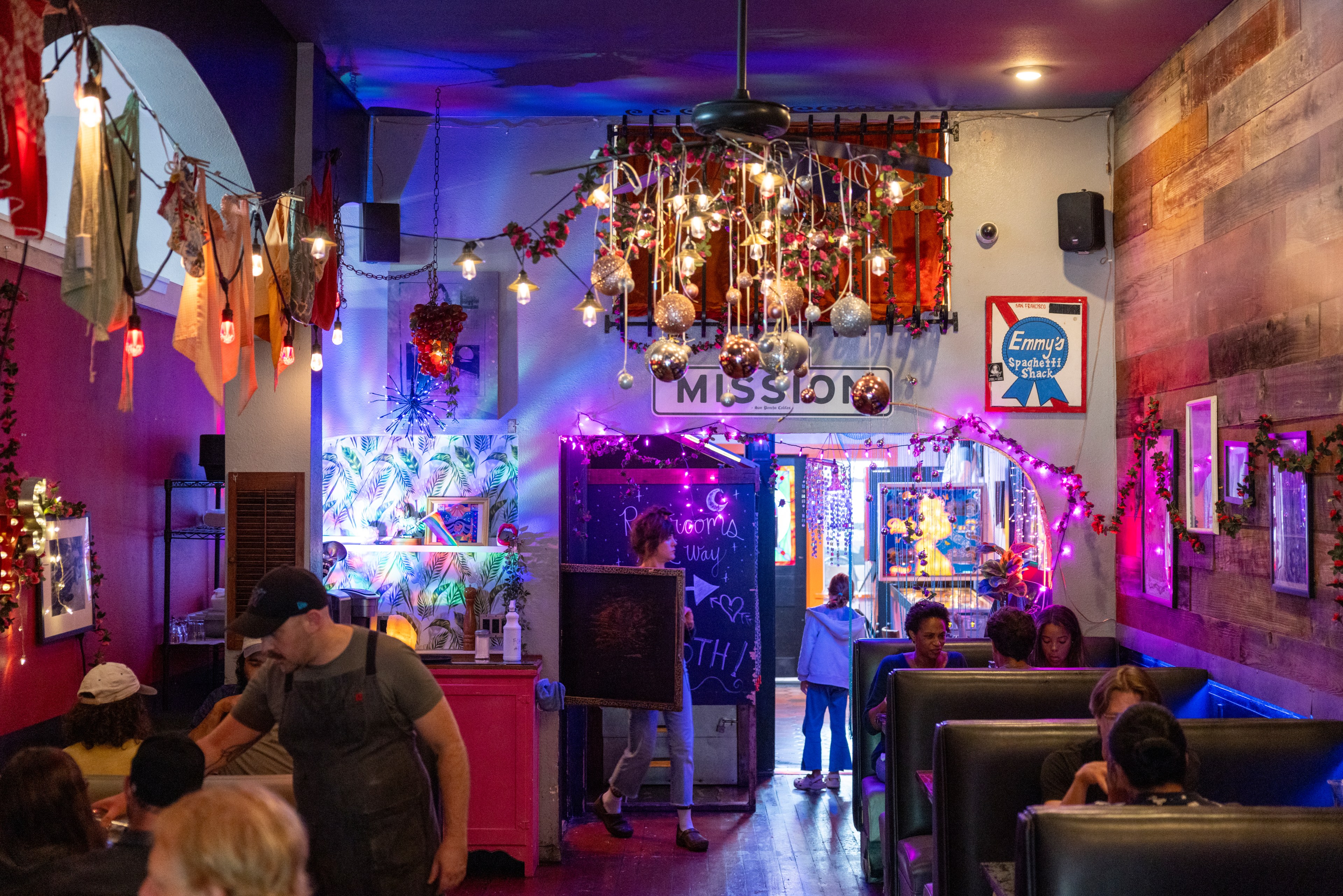 The image shows a cozy, eclectic restaurant with vibrant decorations, string lights, and hanging ornaments. People are seated in booths, and a server is attending to a table.