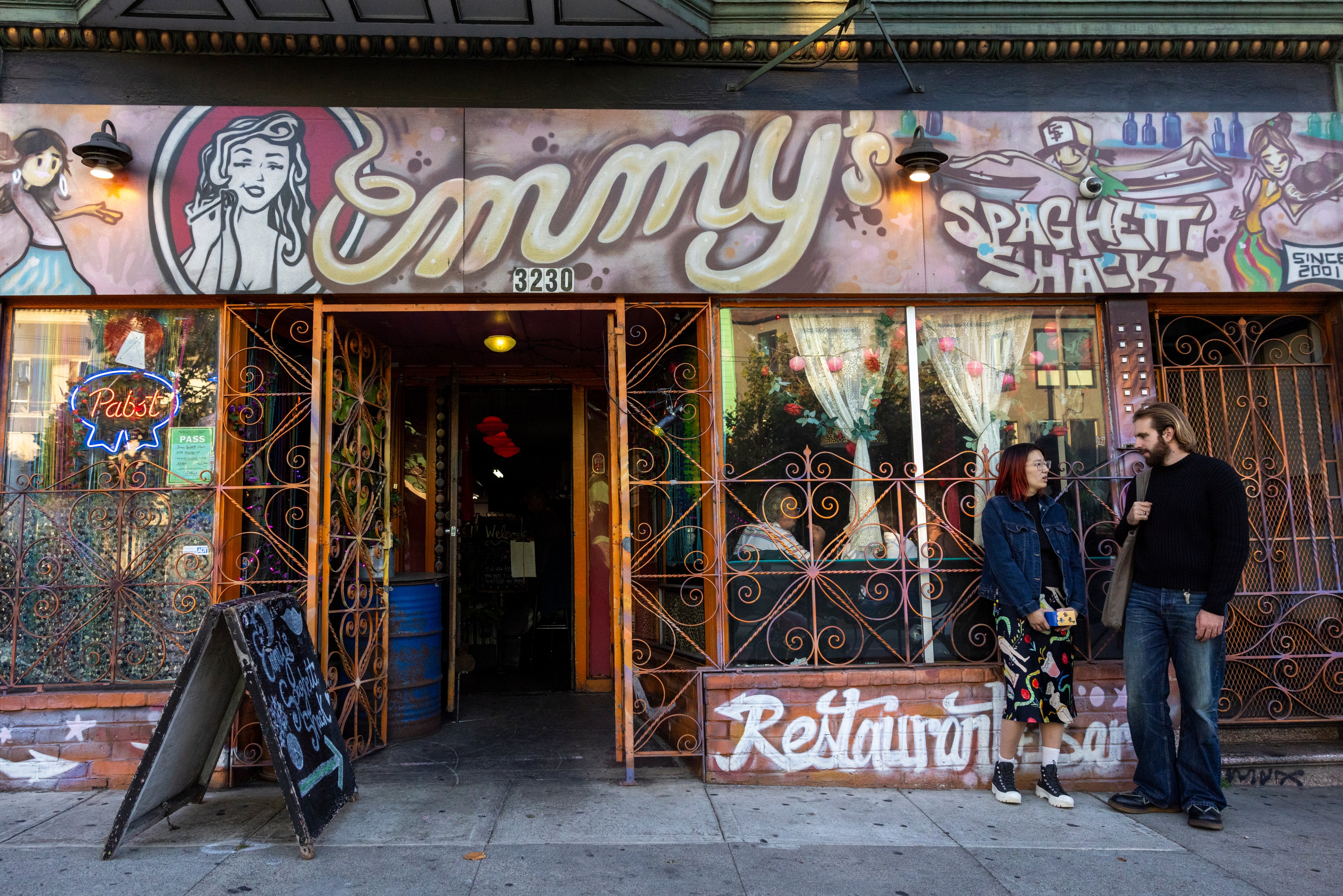 Two people stand outside &quot;Emmy's Spaghetti Shack,&quot; a colorful restaurant with a mural, iron gates, neon signs, and a sidewalk chalkboard sign.