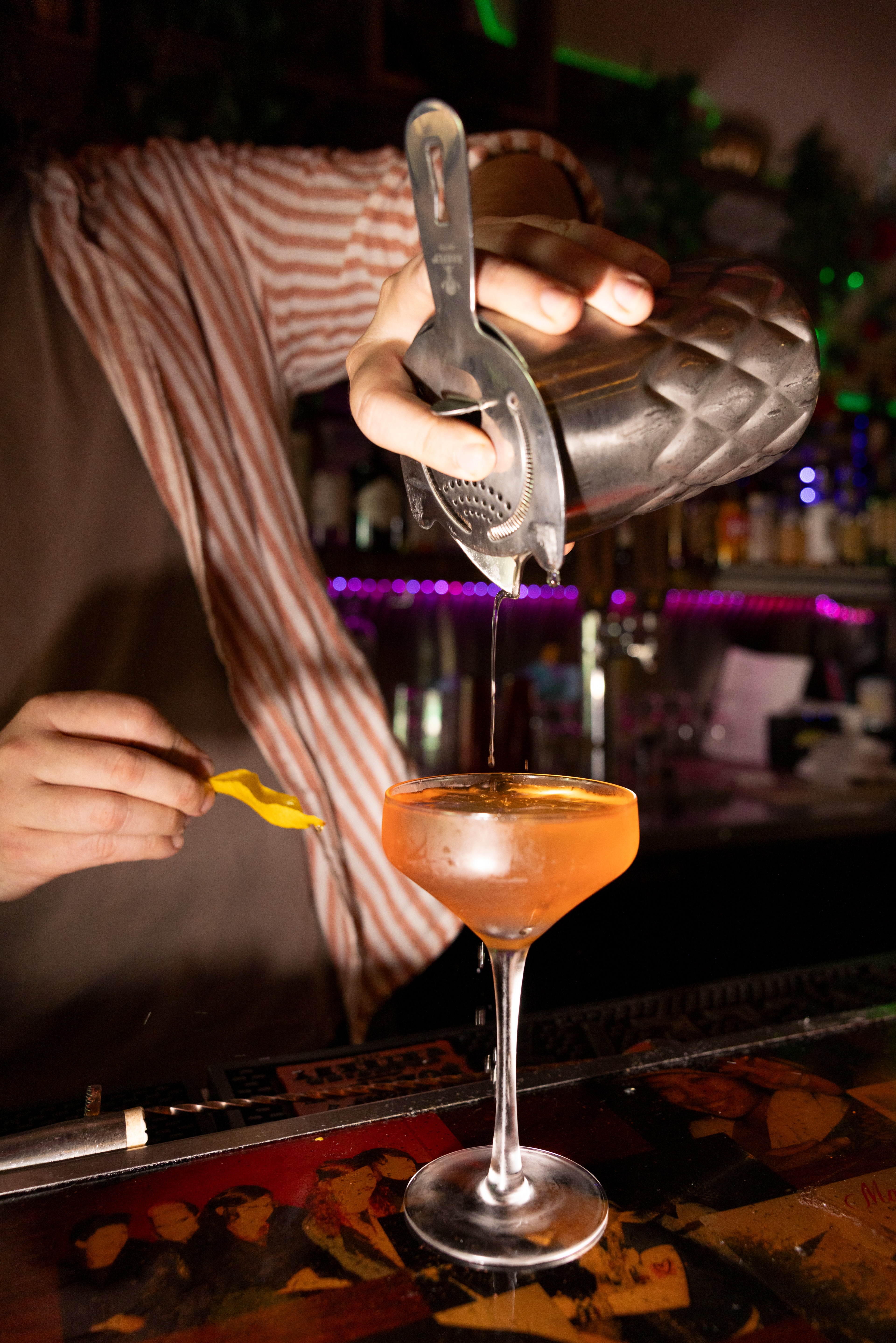 A bartender wearing a striped shirt is pouring a drink from a shaker into a coupe glass, while holding a lemon peel garnish in the other hand.