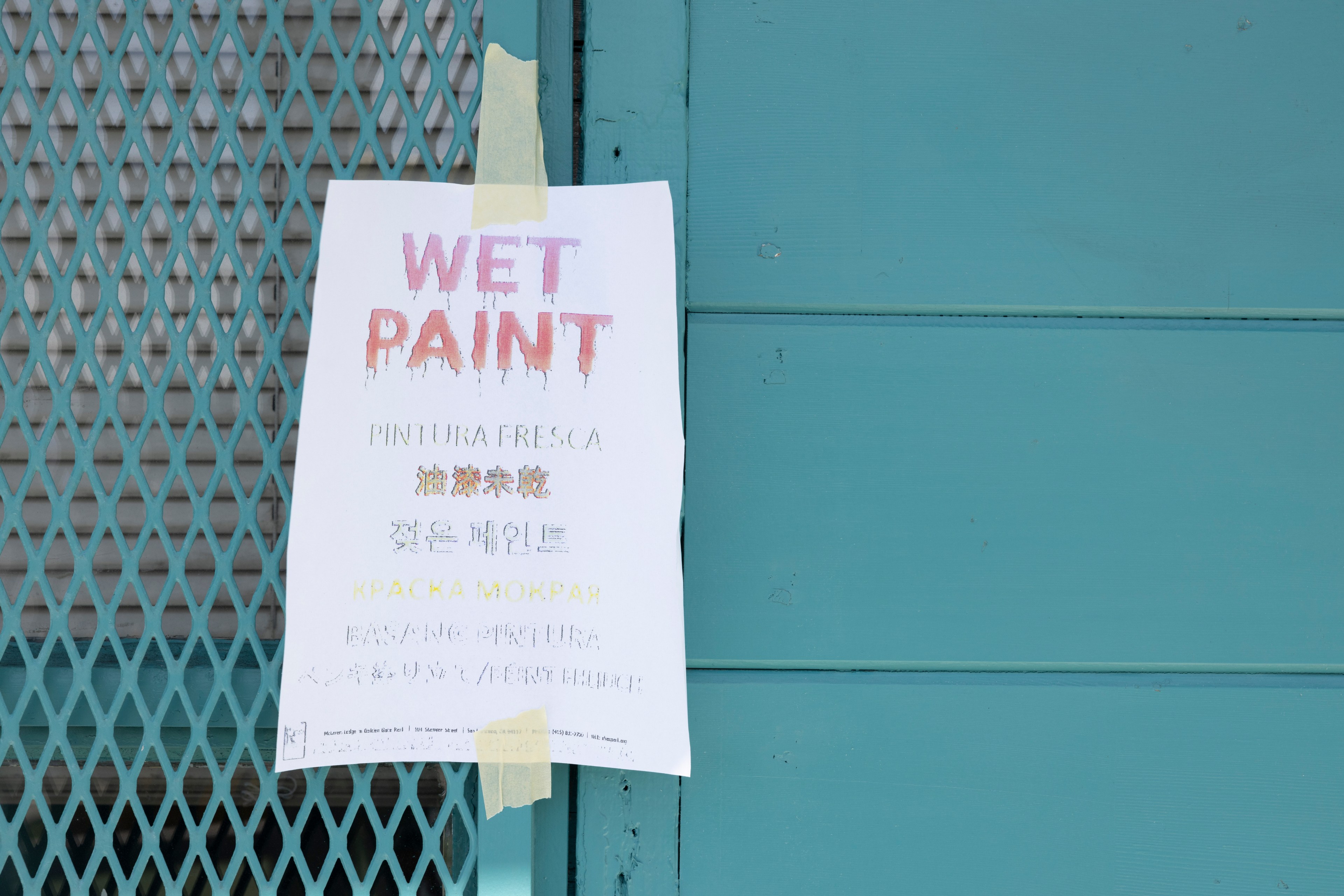 A white poster with &quot;Wet Paint&quot; written in various languages is taped to a turquoise fence and wall, indicating the fresh paint.