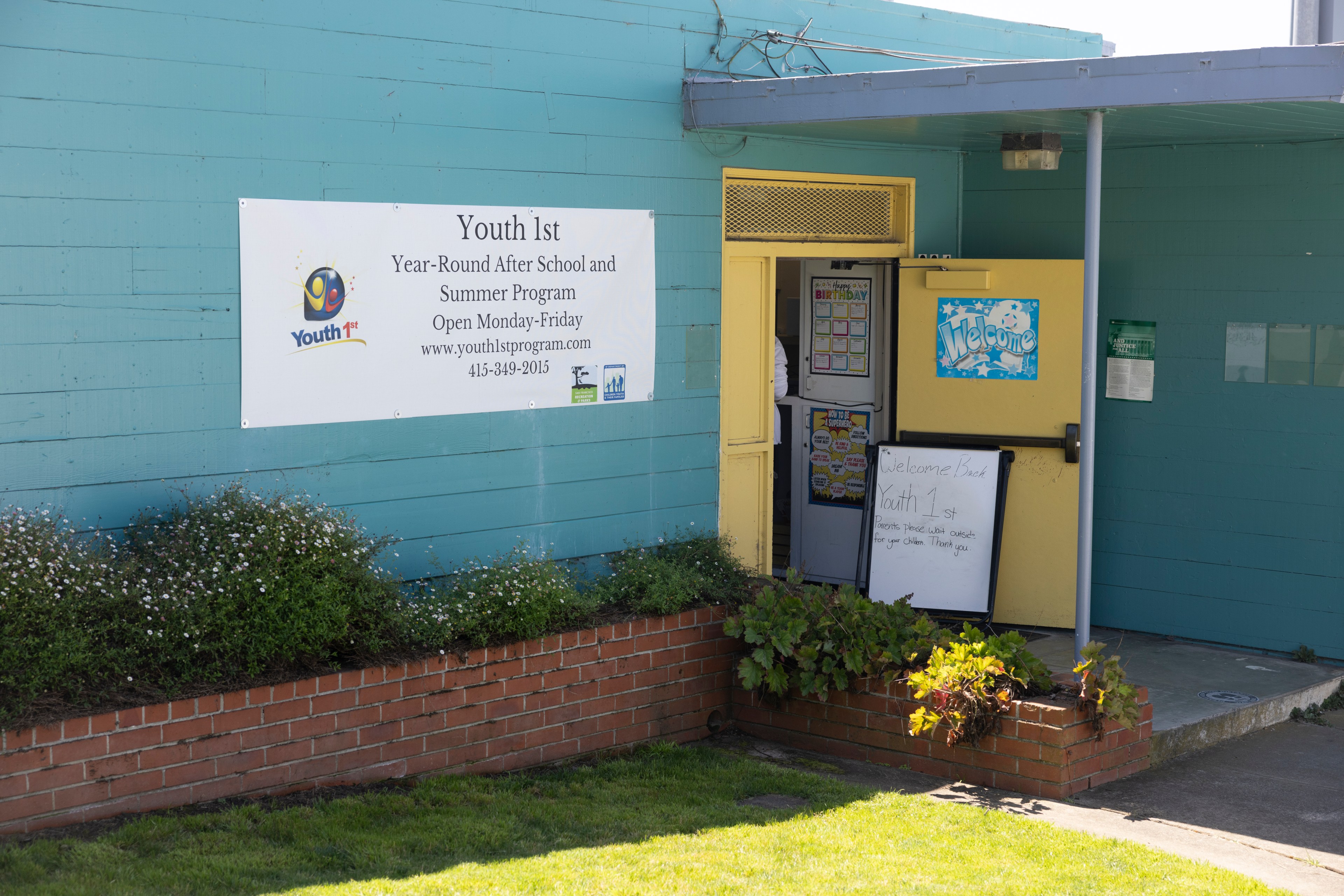 A teal building with a banner for "Youth 1st Year-Round After School and Summer Program" and an open yellow door displaying welcome signs and a whiteboard.