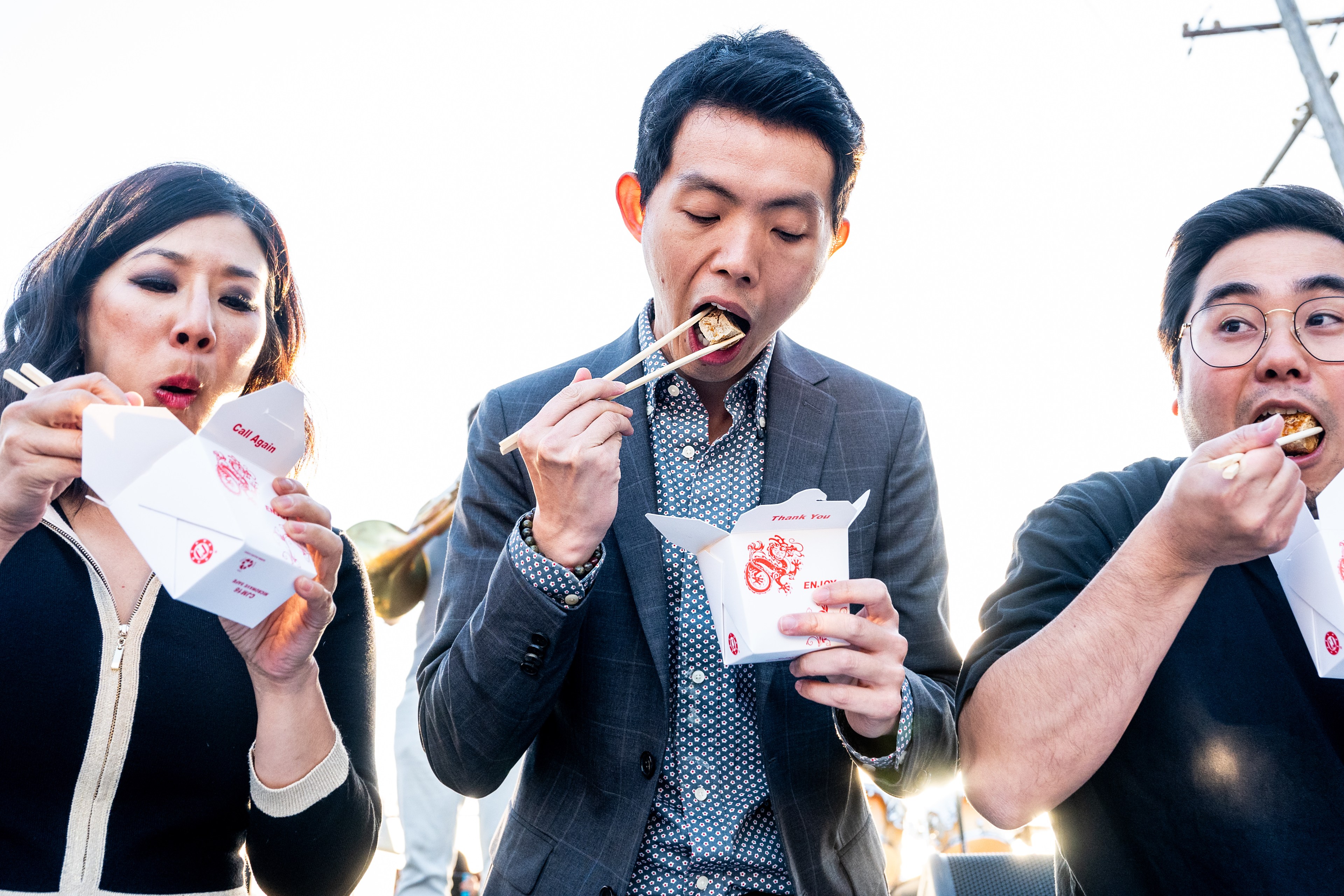 Three people are eating from white takeout boxes using chopsticks; they seem to be enjoying their meal.