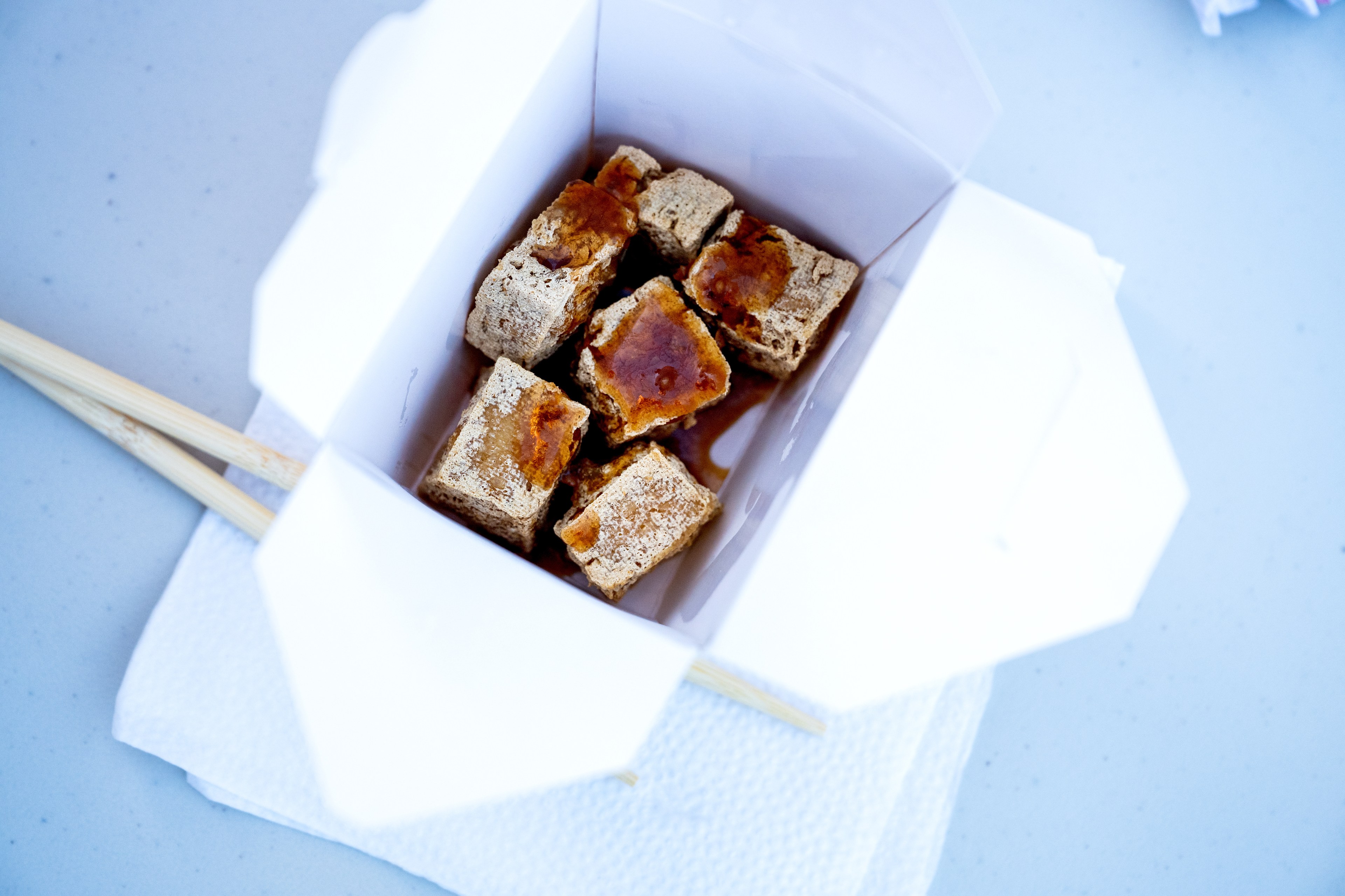 The image shows an opened white takeout box on a napkin containing several cubes of food with a sauce drizzled on top, alongside a pair of wooden chopsticks.