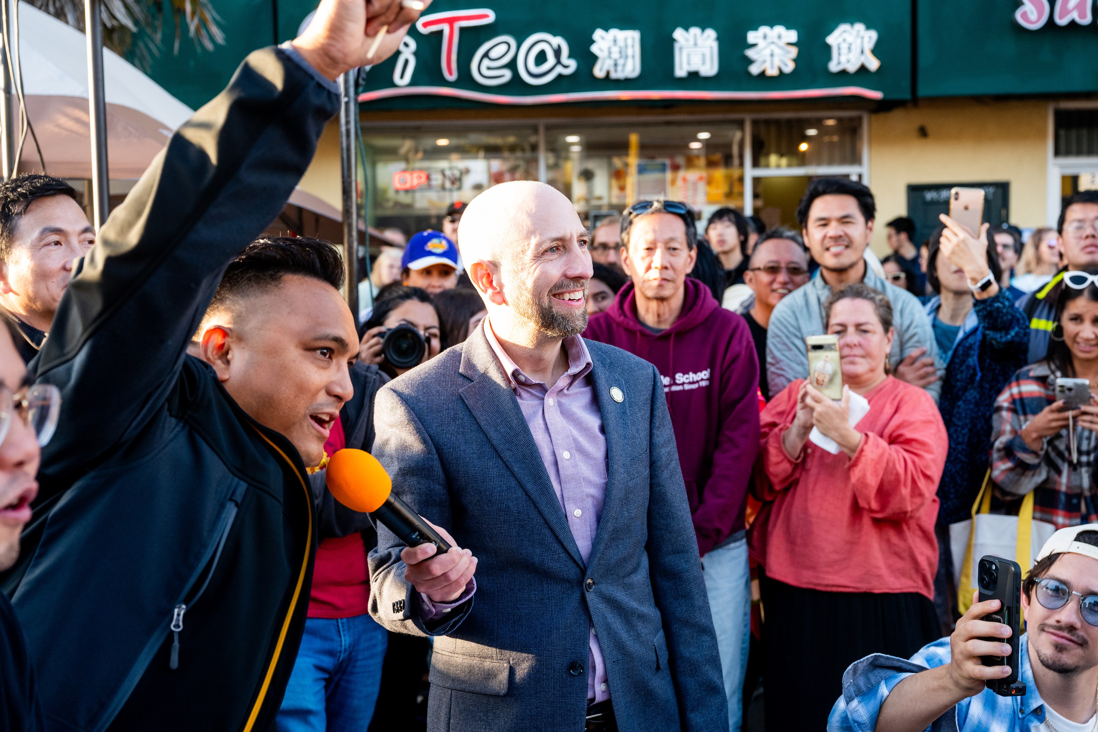 A smiling bald man in a suit stands in a crowd, holding a microphone. Another man gestures energetically, with people around them taking photos.