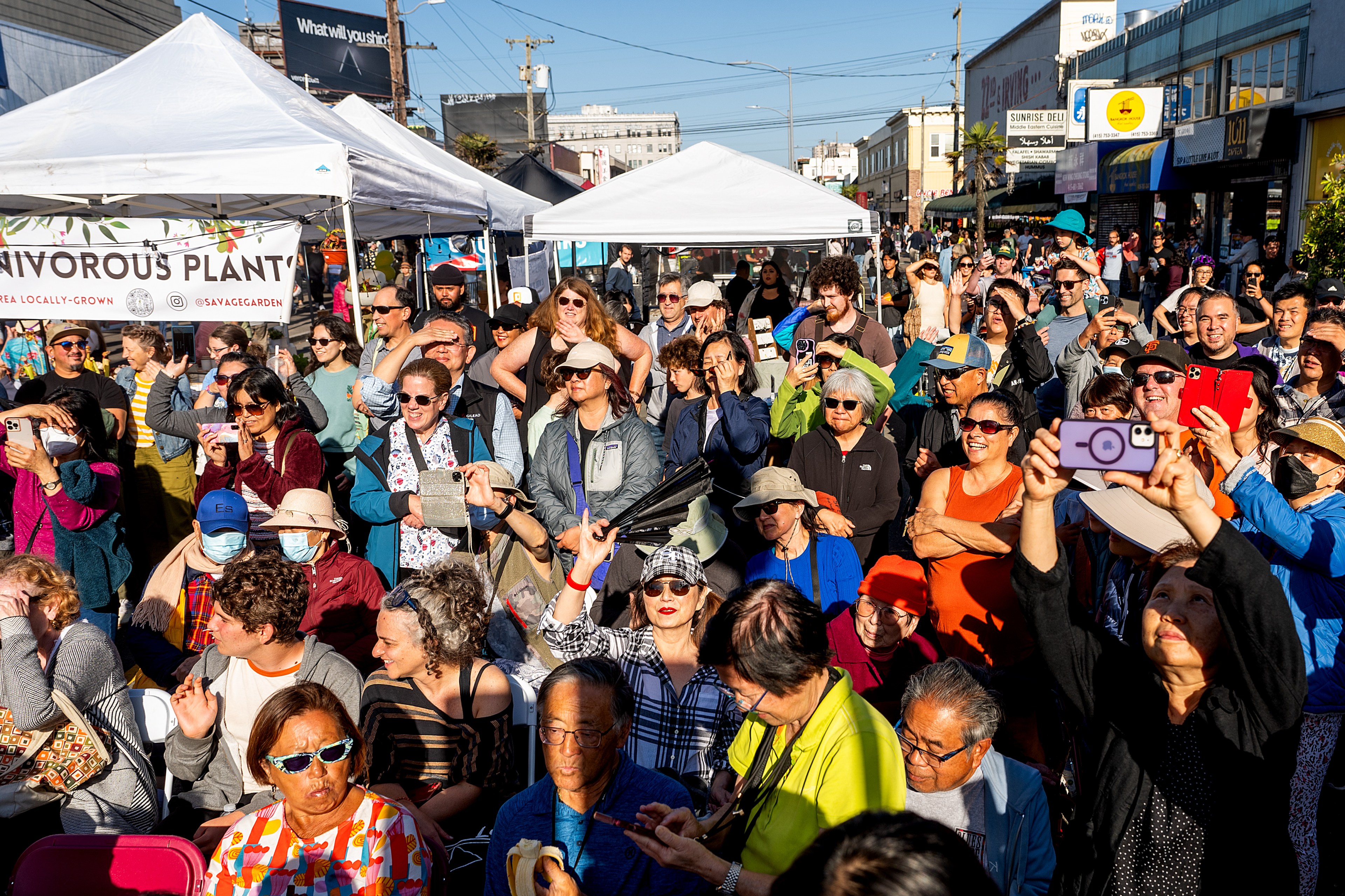 A lively crowd is gathered outdoors under white tents. People are smiling, raising their hands, and taking photos with smartphones. The scene is colorful and bustling.