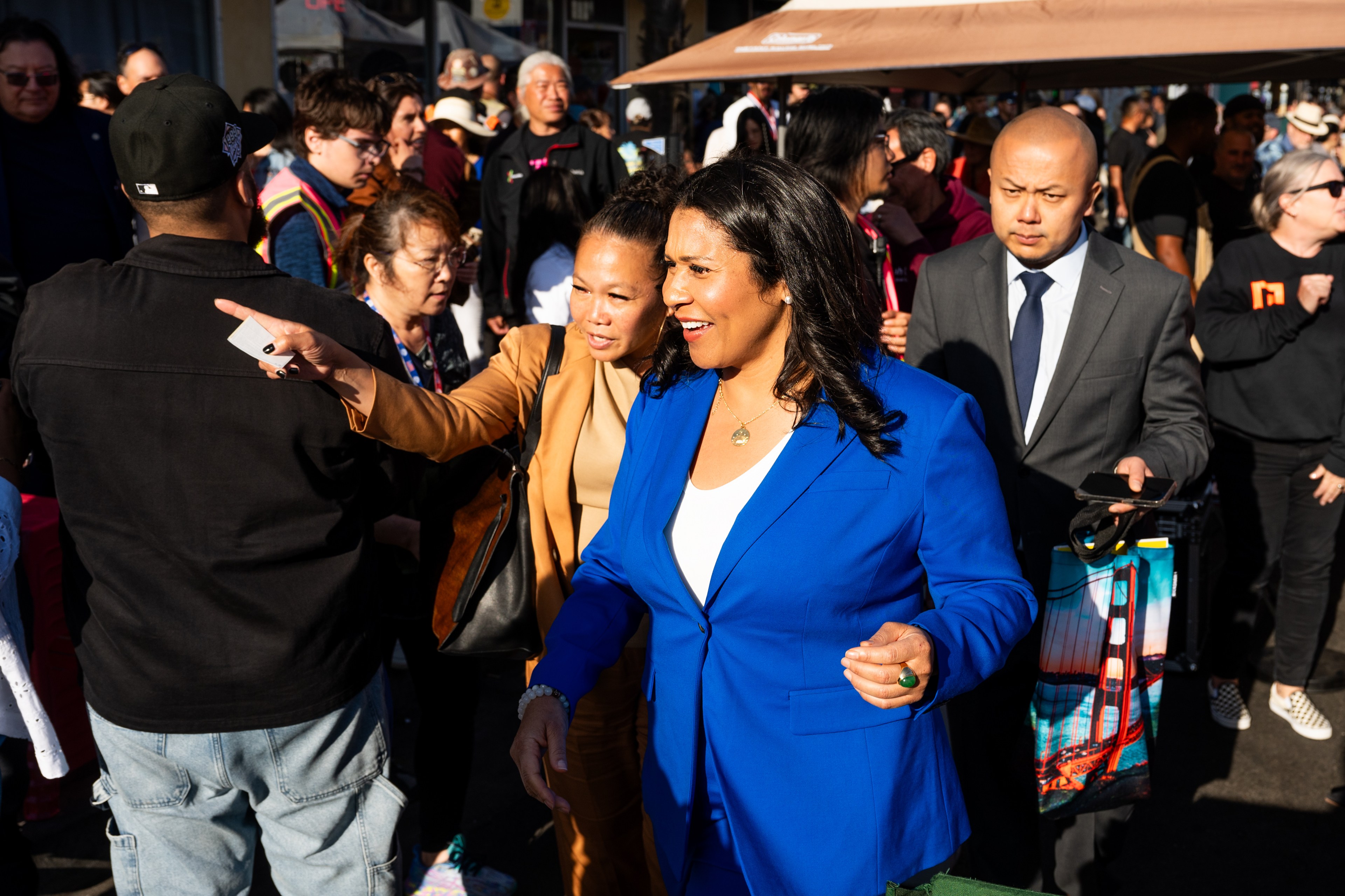 A woman in a bright blue suit walks in a busy, sunny outdoor crowd, smiling. A woman points and another man in a suit holds a smartphone and a colorful bag.