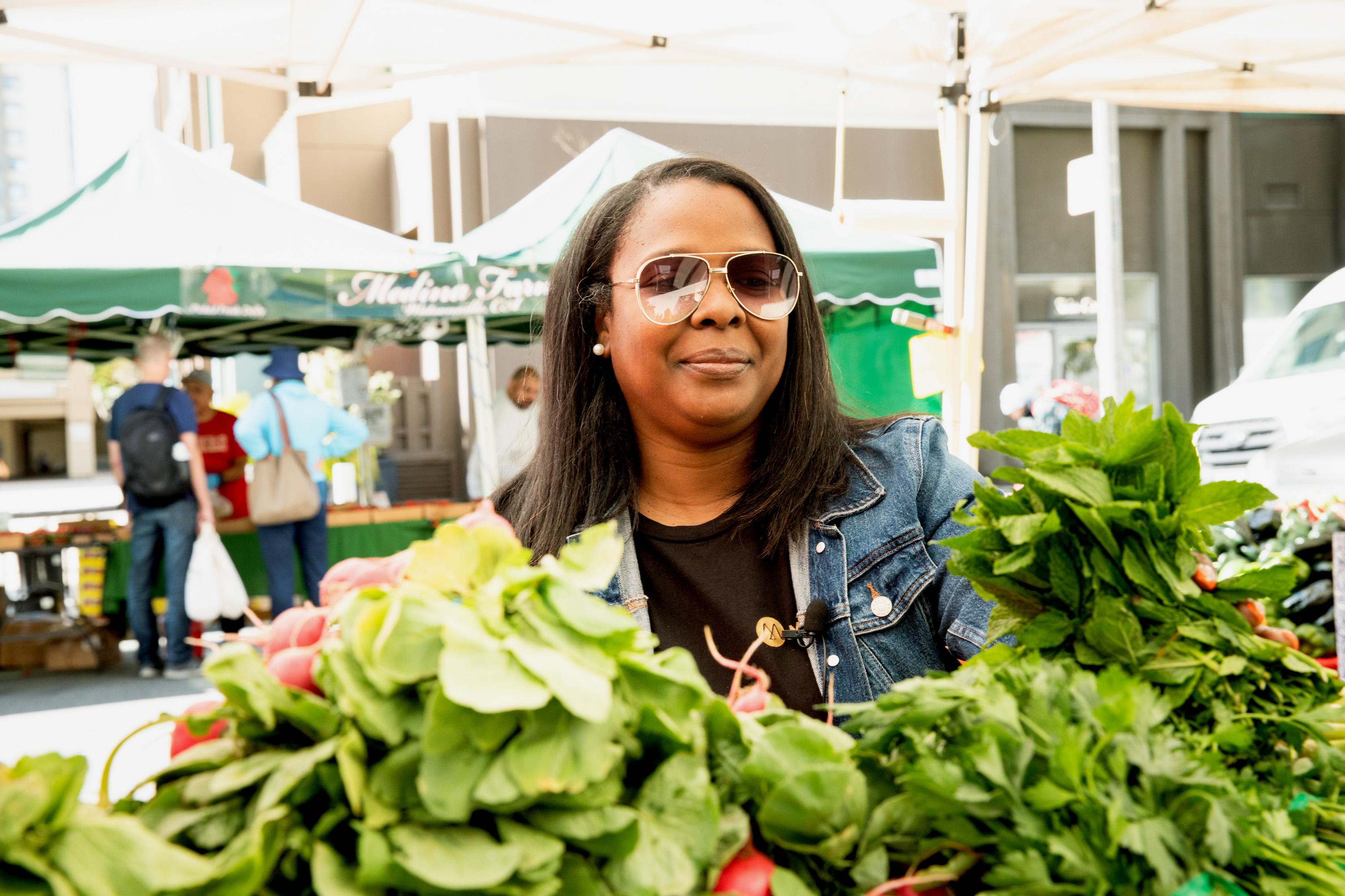 A woman in sunglasses and a denim jacket stands behind a display of fresh greens at an outdoor market with green tents and people in the background.