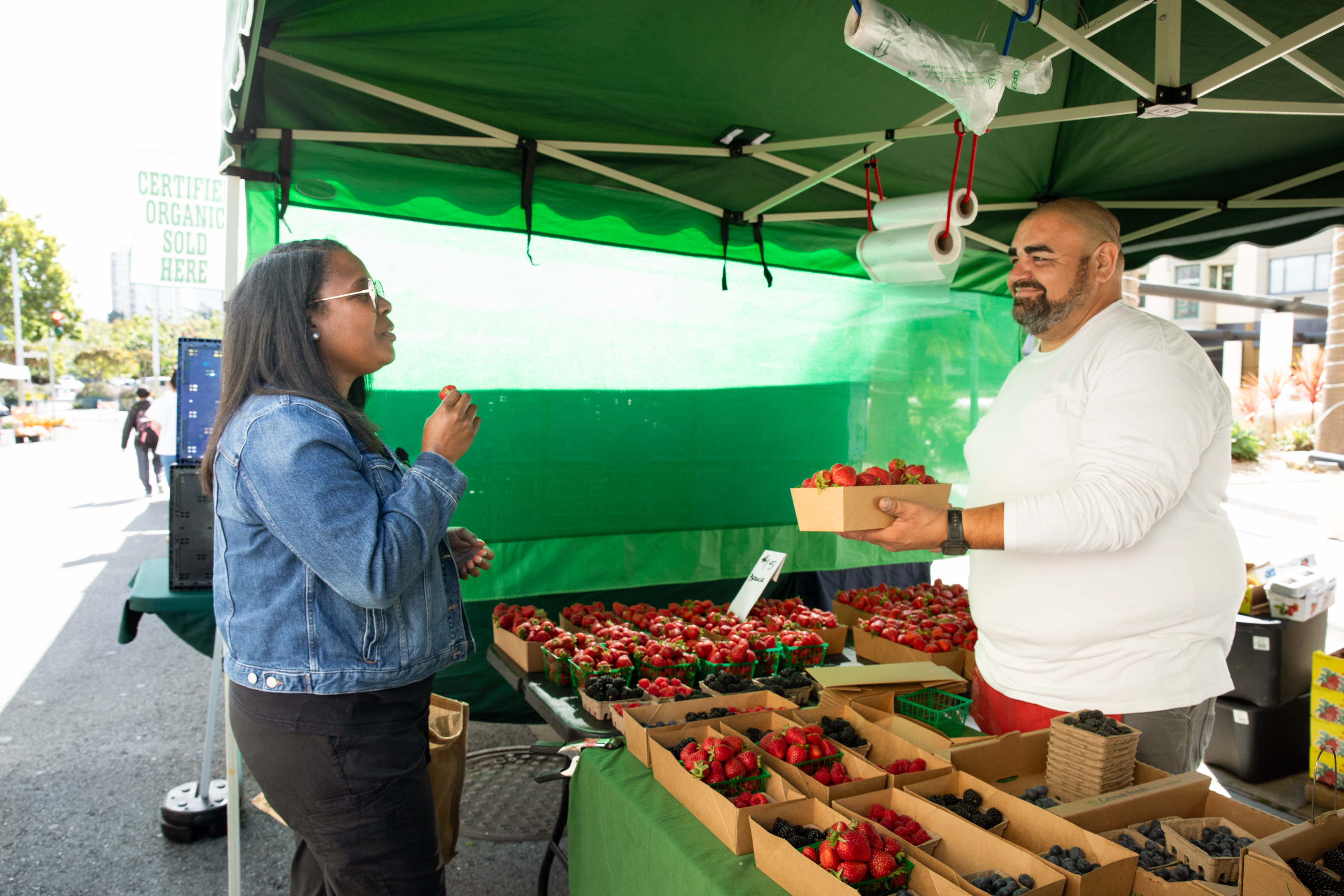 A woman in a denim jacket is sampling strawberries at a farmer's market stand, while a man in a white shirt holds a container of strawberries under a green tent with more berries on display.