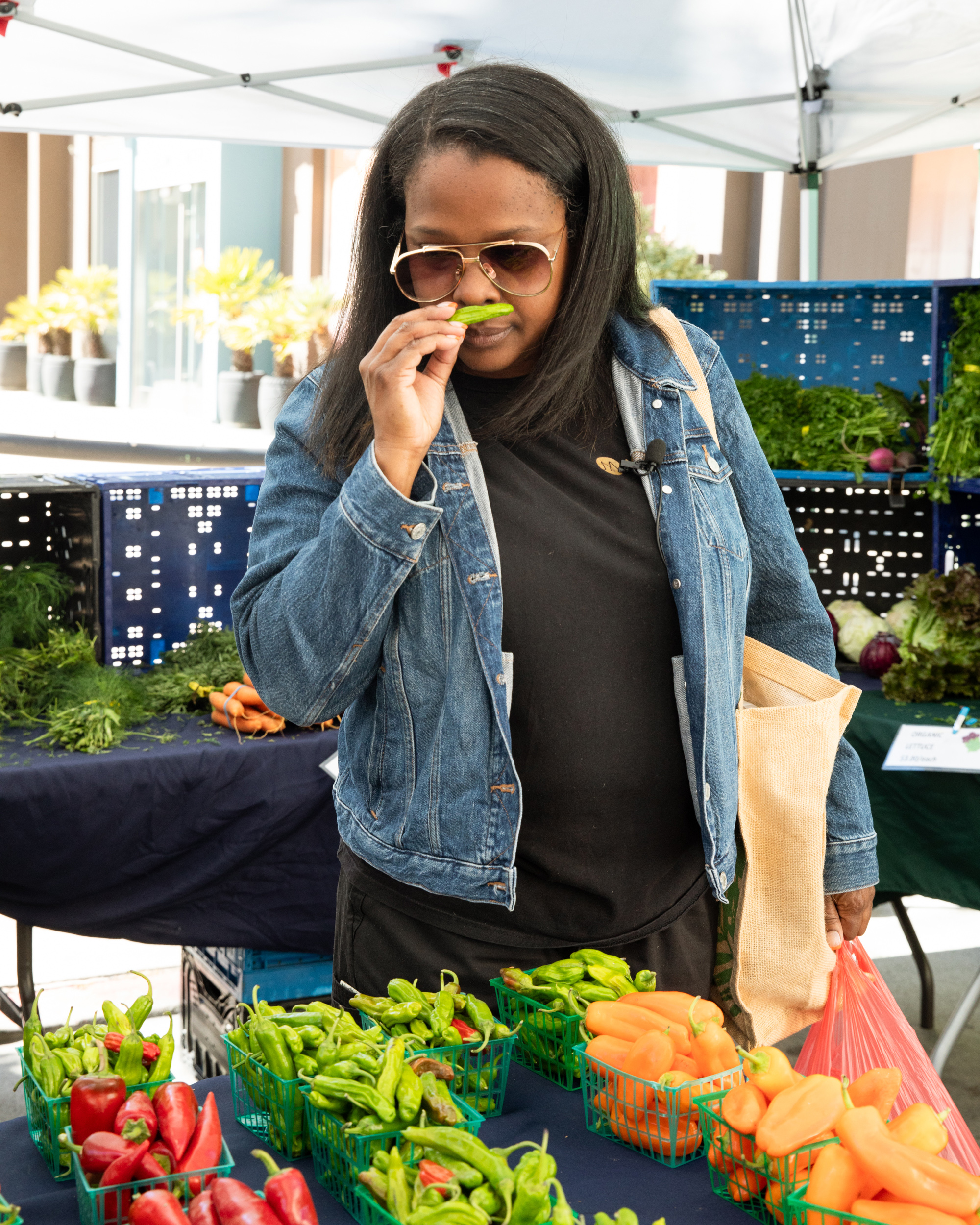 A woman in sunglasses and a denim jacket smells a green pepper at an outdoor market surrounded by various fresh vegetables like red and green peppers.