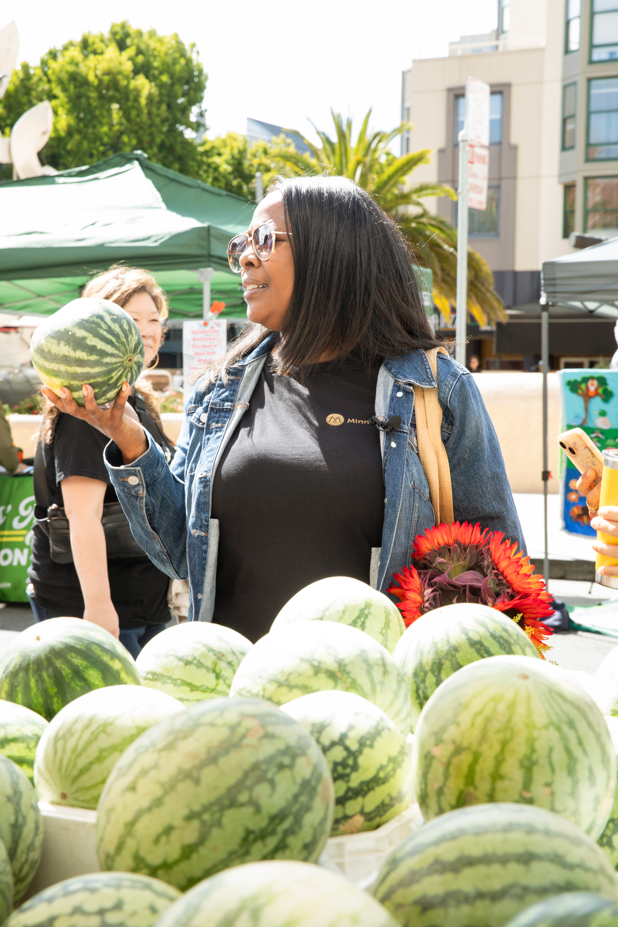 A woman is holding a watermelon at an outdoor market. She wears sunglasses and a denim jacket, with another watermelon in front of her and sunflowers in her tote bag.