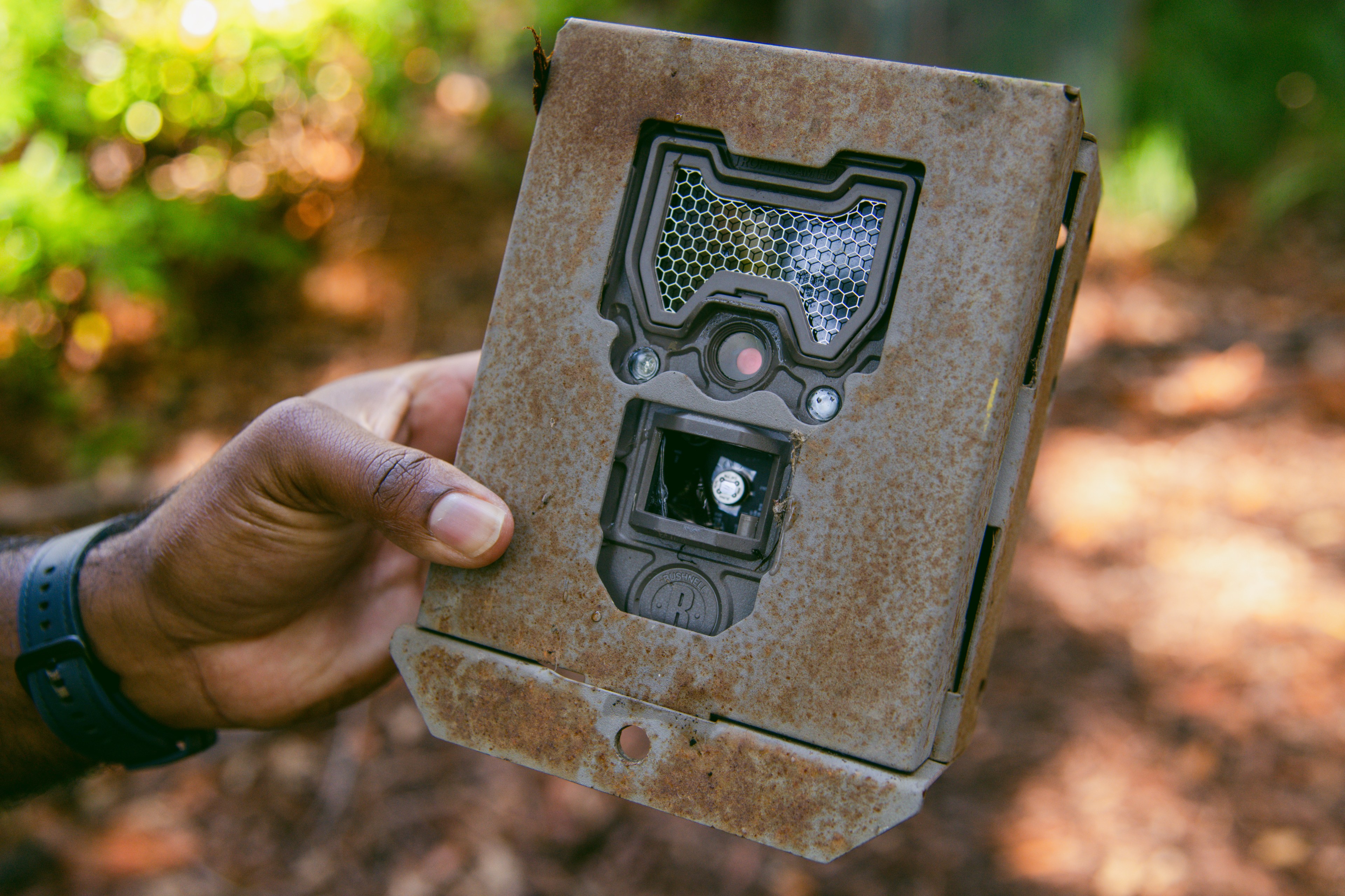 A hand holds an old, rusted rectangular device with a mesh-covered area, a lens, and an open slot. The background is a blurred outdoor setting with greenery.