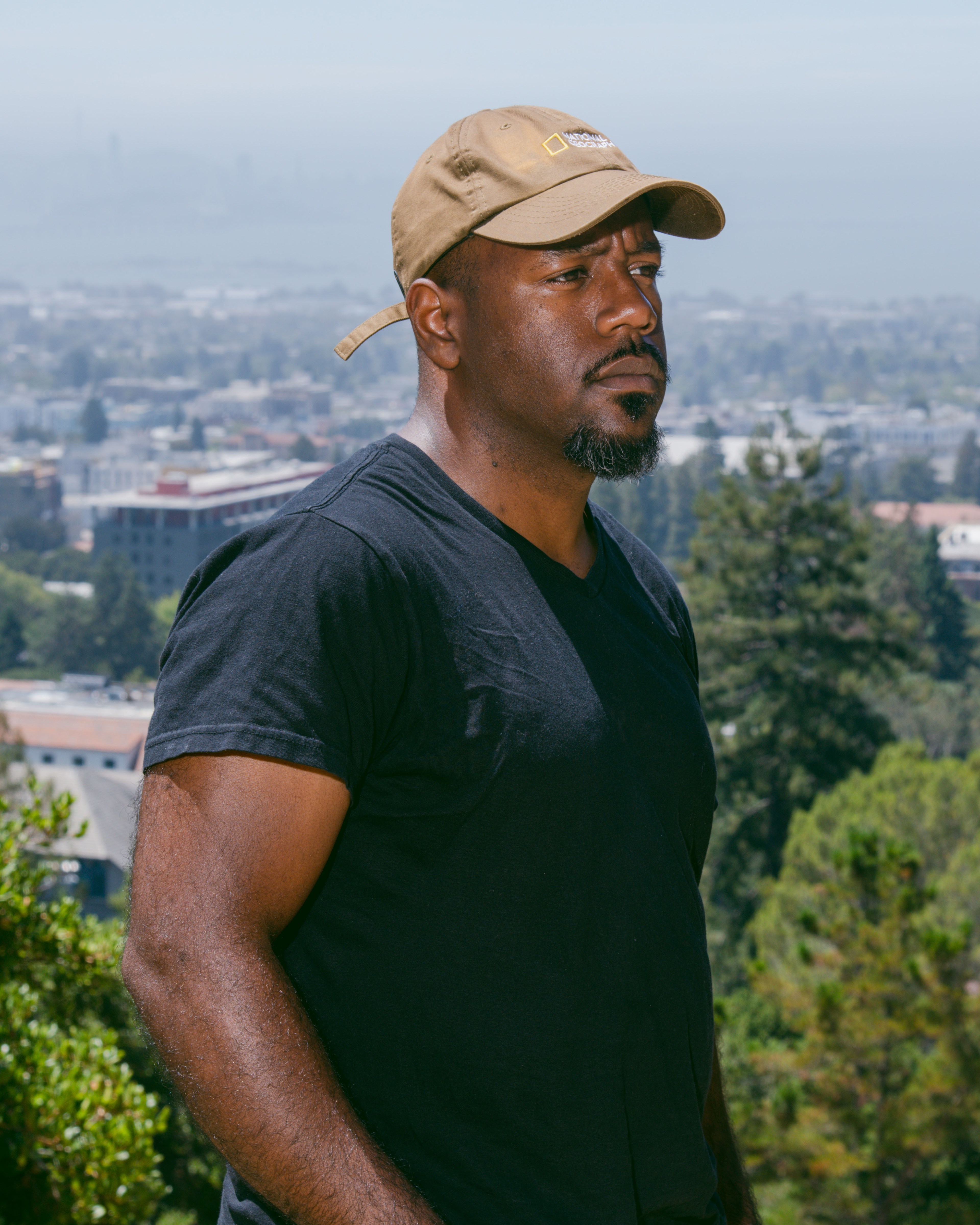 A man wearing a brown cap and black t-shirt stands outdoors, looking serious. Behind him is a blurred view of a city with trees and buildings.