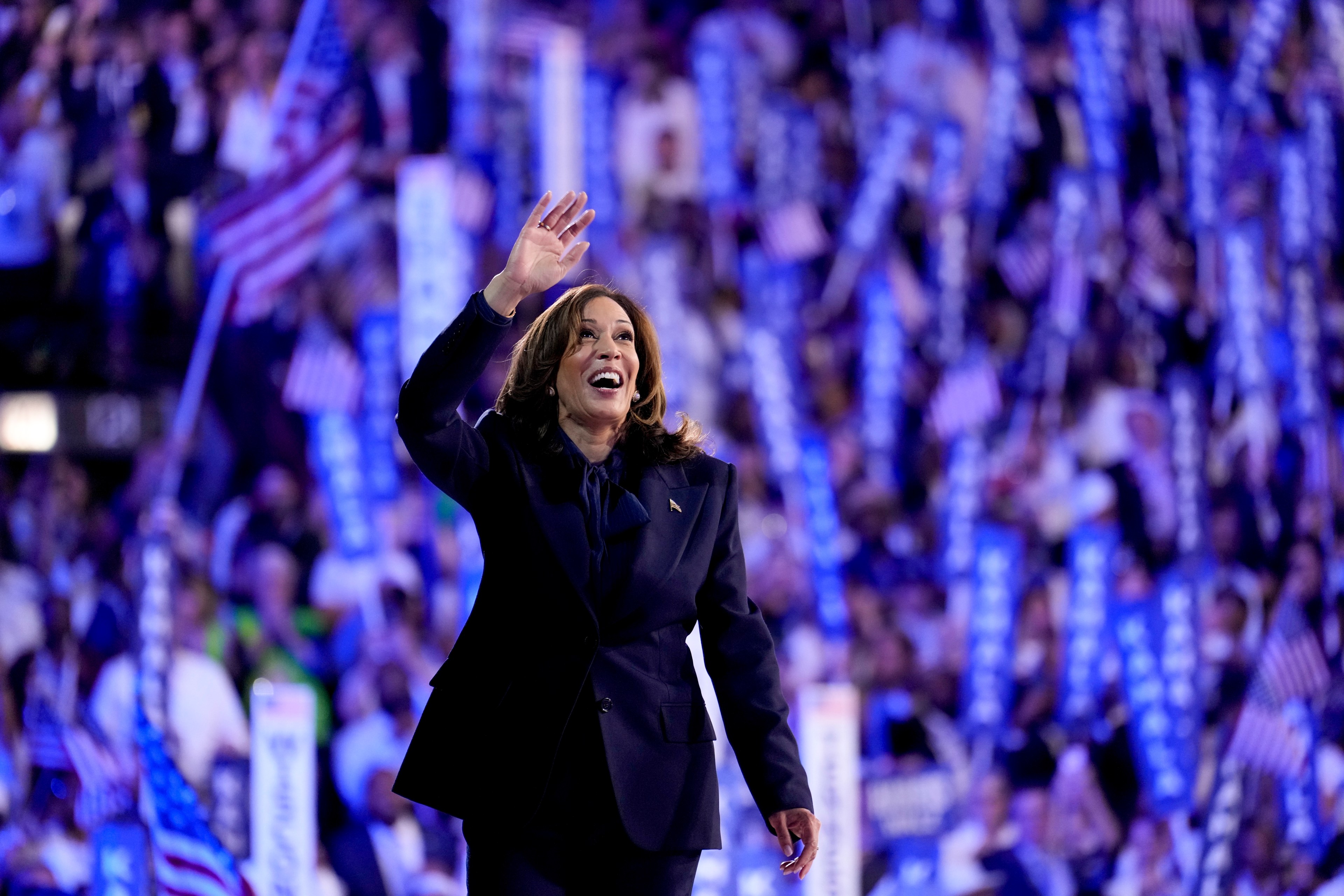 A woman in a dark suit waves and smiles, standing in front of a large, cheering crowd with American flags and blue banners in the background.