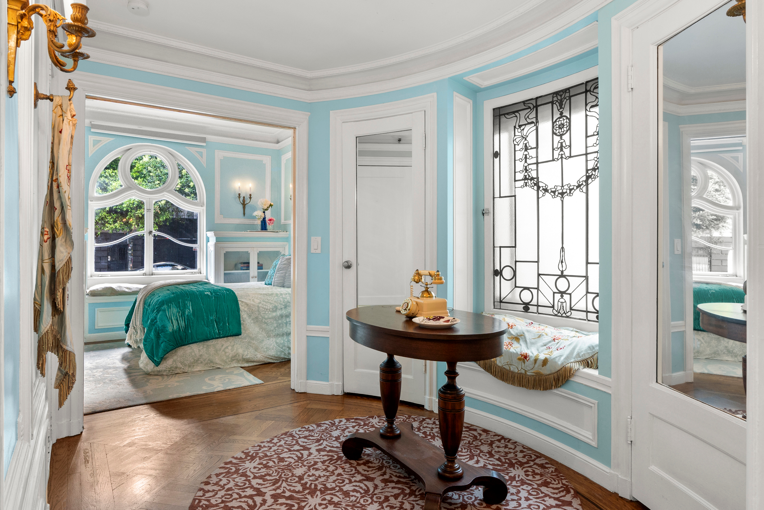 A light blue room with ornate white trim features a bed with a green cover, a round window, an arched window, and a small sitting area with an antique phone.