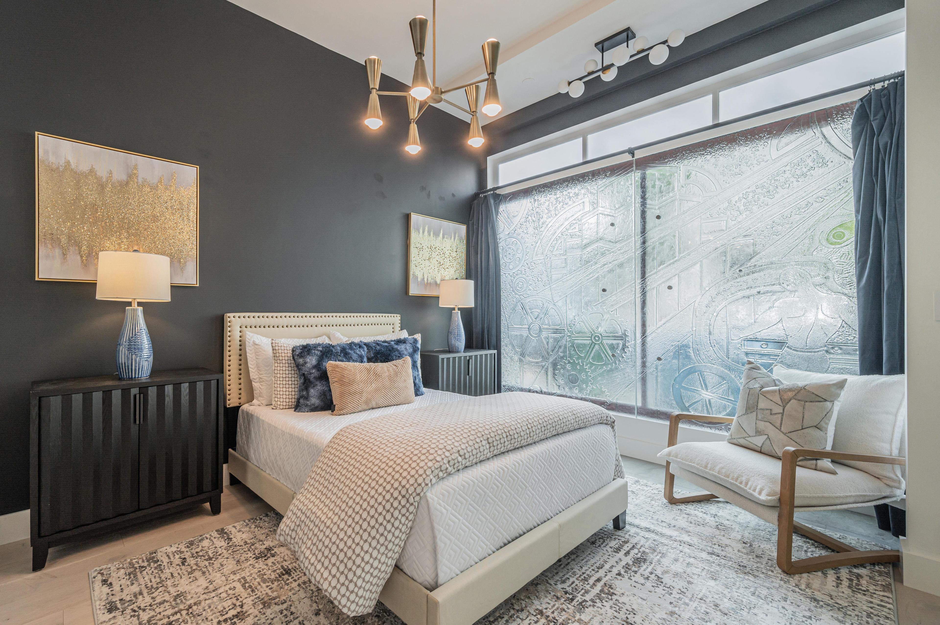 A modern bedroom features a bed with white bedding, dark accent wall, two side tables with lamps, abstract wall art, a patterned rug, and large frosted windows.