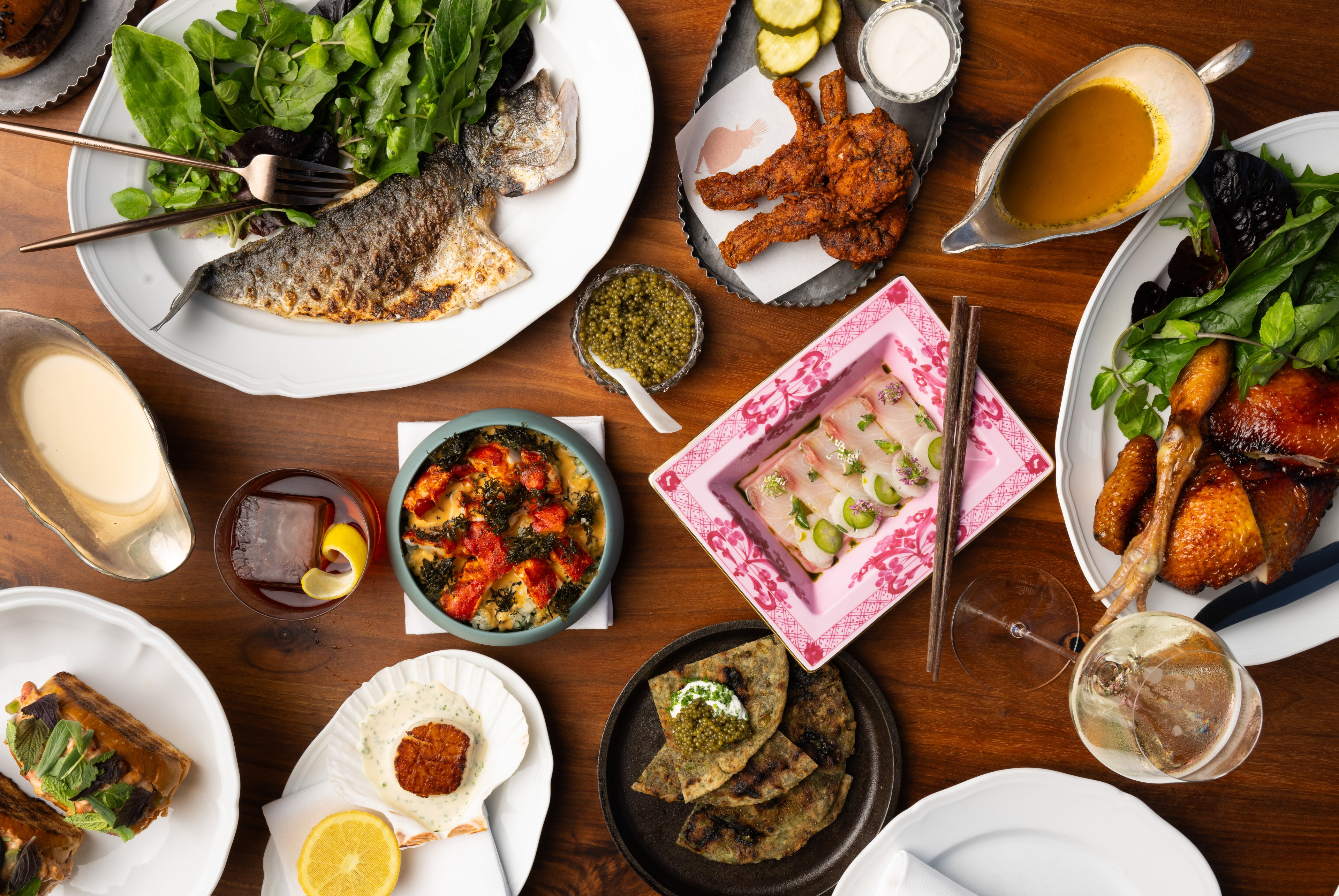 A variety of dishes are displayed on a wooden table including fish, fried chicken, salad, a cocktail, sauces, and garnished vegetables.