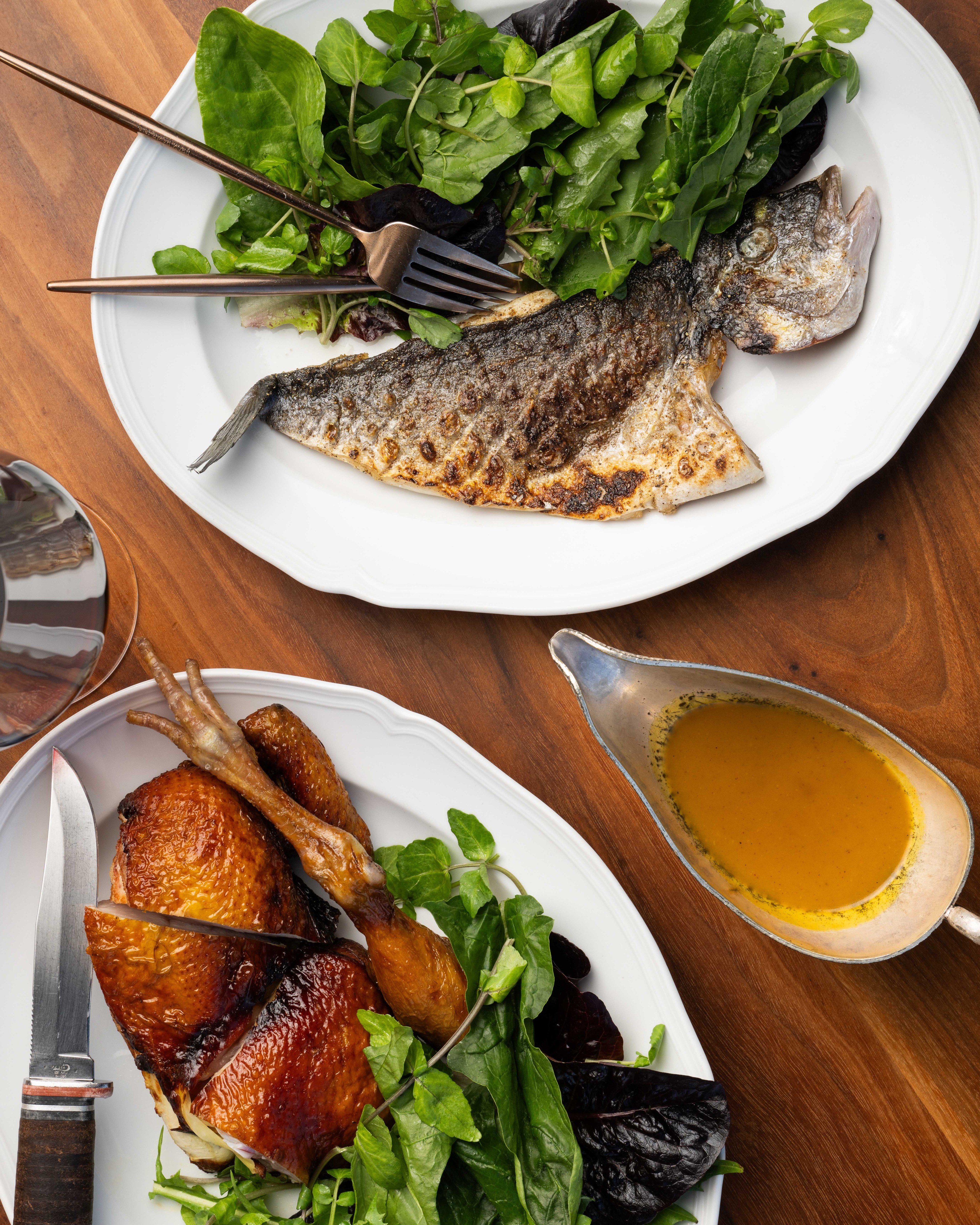 The image shows a grilled fish with leafy greens on one plate and roasted chicken with greens on another, alongside a gravy boat with golden sauce, all on a wooden table.