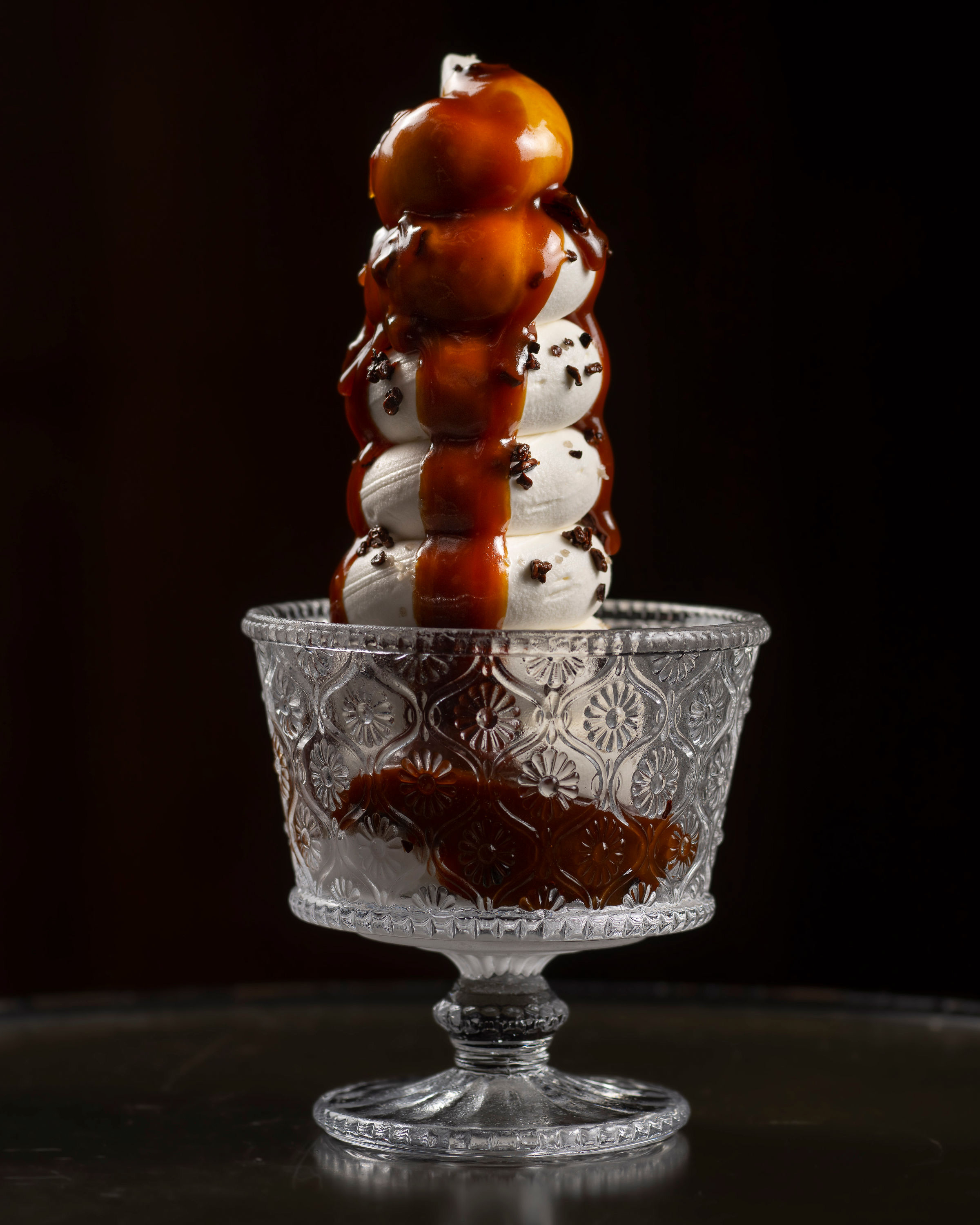 A tall stack of white meringue kisses in a patterned, glass dish, covered in rich caramel sauce, with dark sprinkles and a dark background.