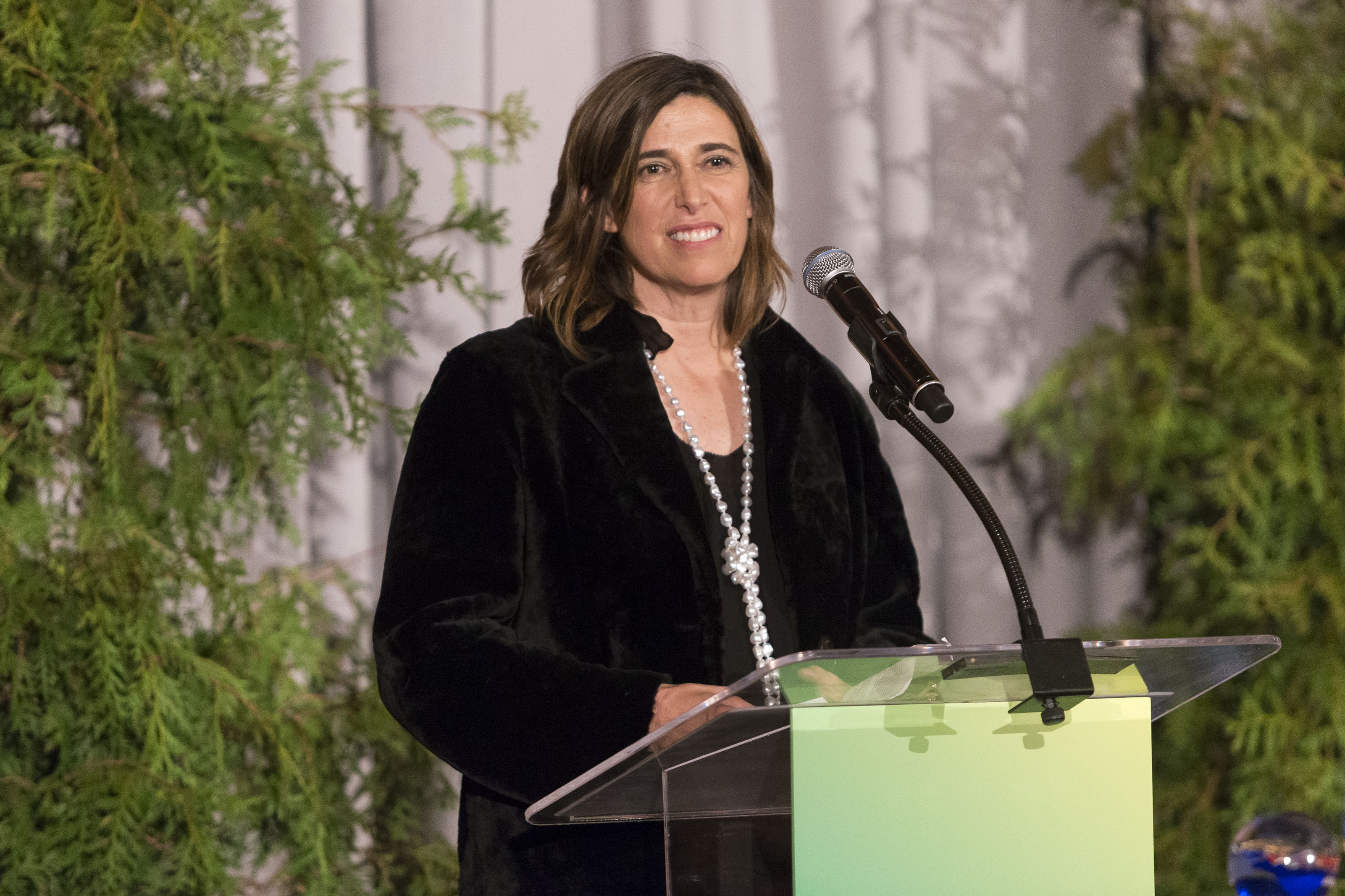 A woman in a black jacket stands at a podium with a microphone, speaking. Green foliage is in the background. She is smiling and has shoulder-length brown hair.