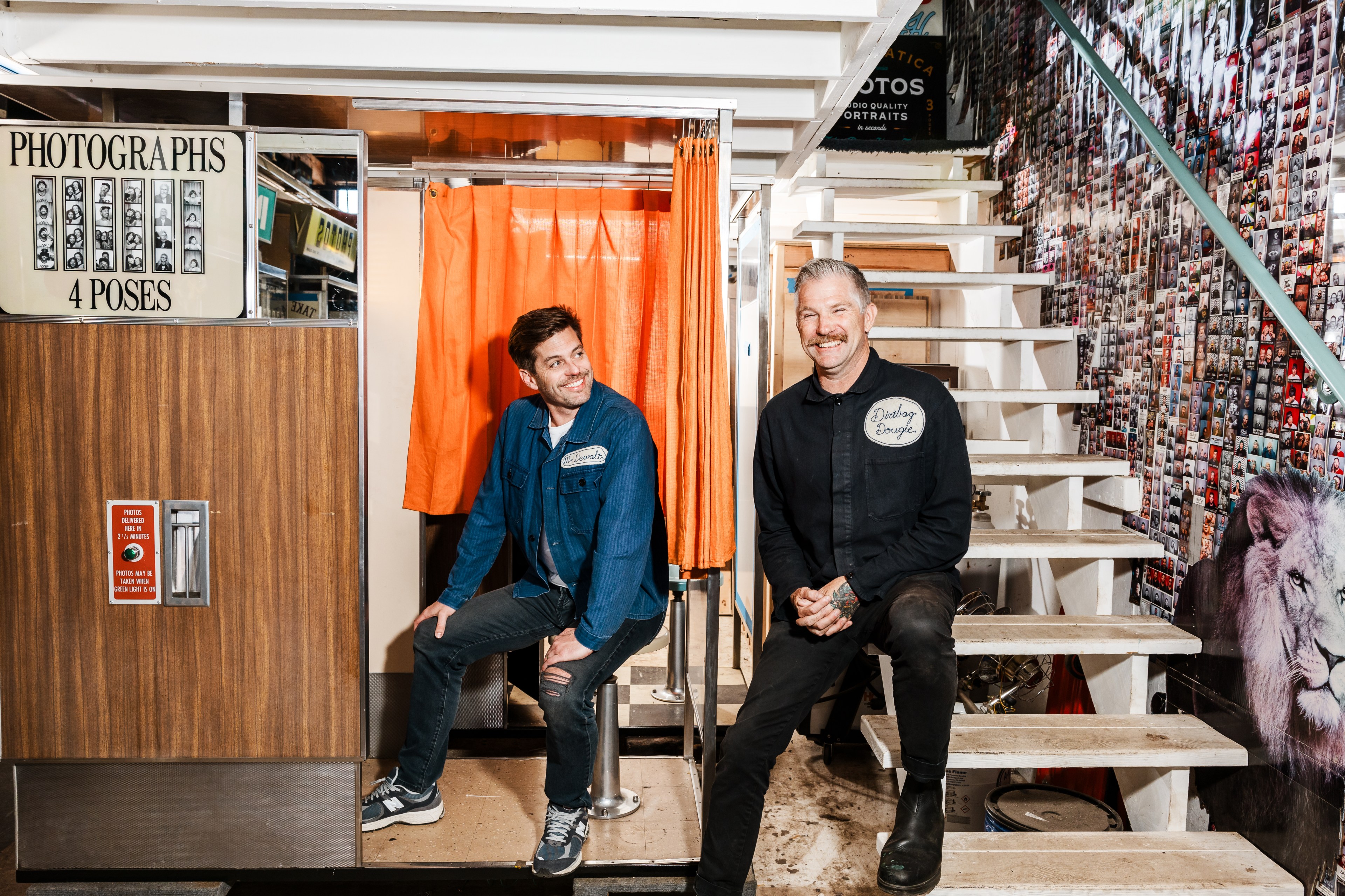 Two men are smiling, one sits in a photo booth with orange curtains, the other sits on stairs covered with photos. The booth sign reads &quot;Photographs, 4 Poses&quot;.