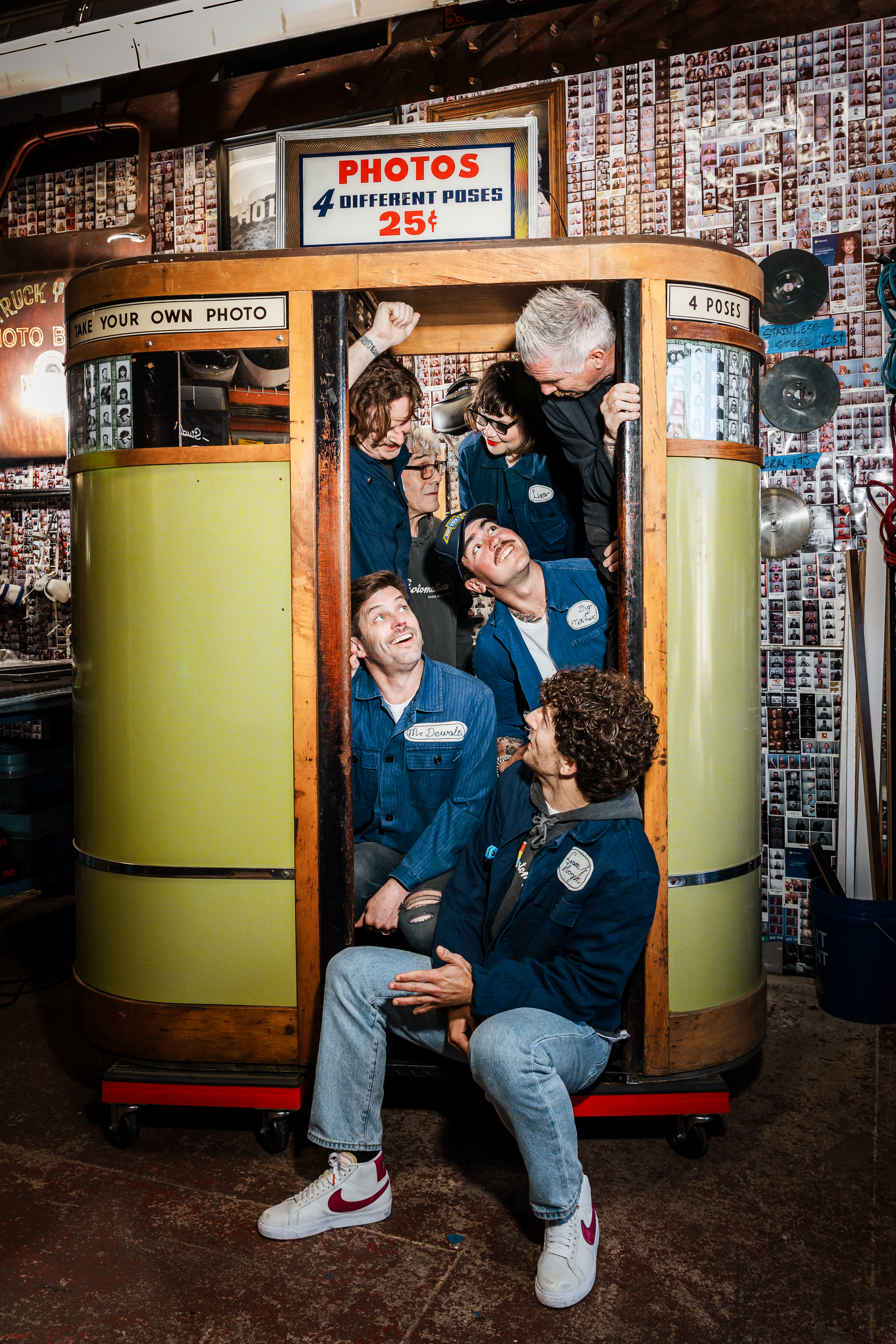 Six people in denim uniforms are crammed inside a vintage photo booth, laughing and looking at each other. Polaroid photos cover the walls around them.