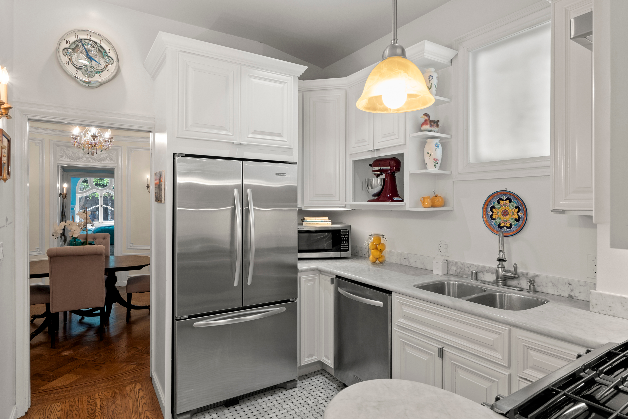 A modern kitchen with white cabinets, stainless steel appliances, and a marble countertop. Bright pendant light, wall clock, and adjoining dining room visible.