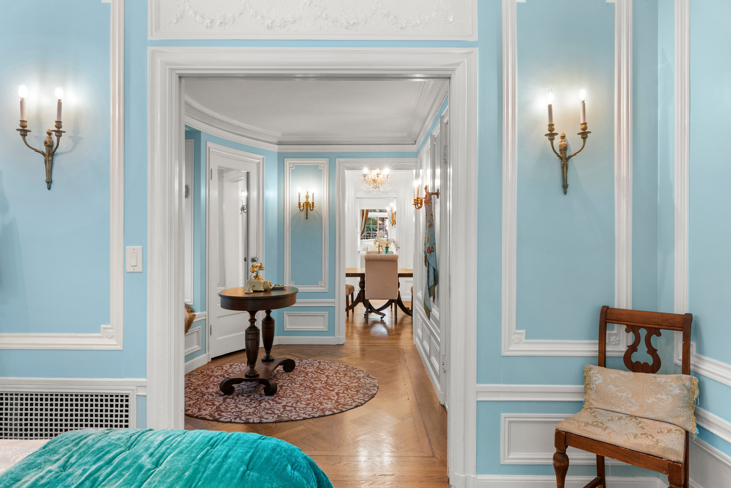 The image shows an elegant room with light blue walls and white trim, featuring classic wall sconces, a small round table with decor, a wooden chair, and views into an adjacent area.