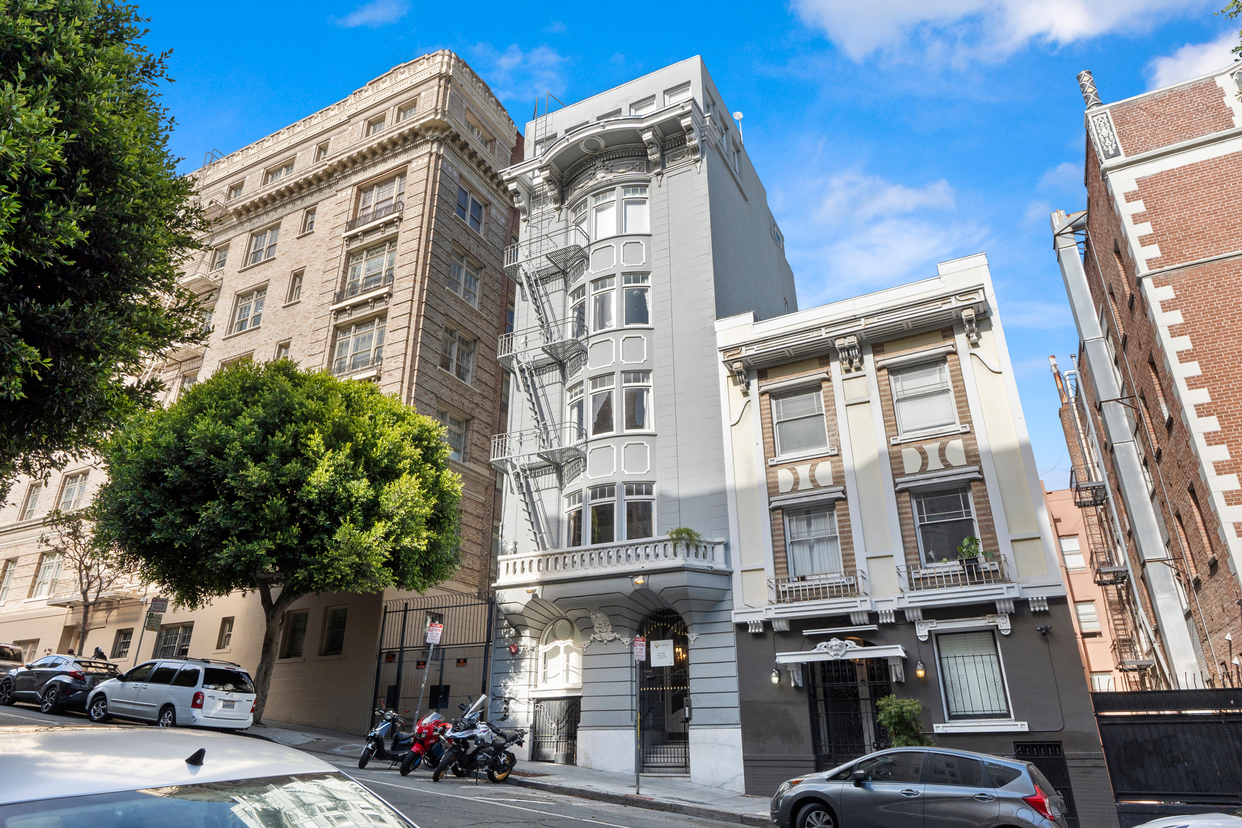 The image shows a street with several multi-story buildings displaying varied architectural designs. There are parked cars, green trees, and a blue sky.