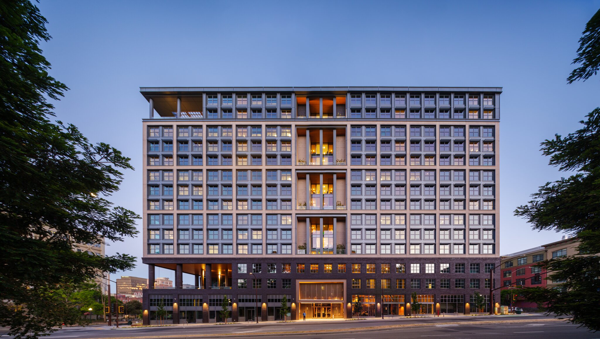 The image shows a modern, multi-story building with a grid-like window pattern and a mix of light and dark facade elements, set against a clear sky. Trees frame the sides.