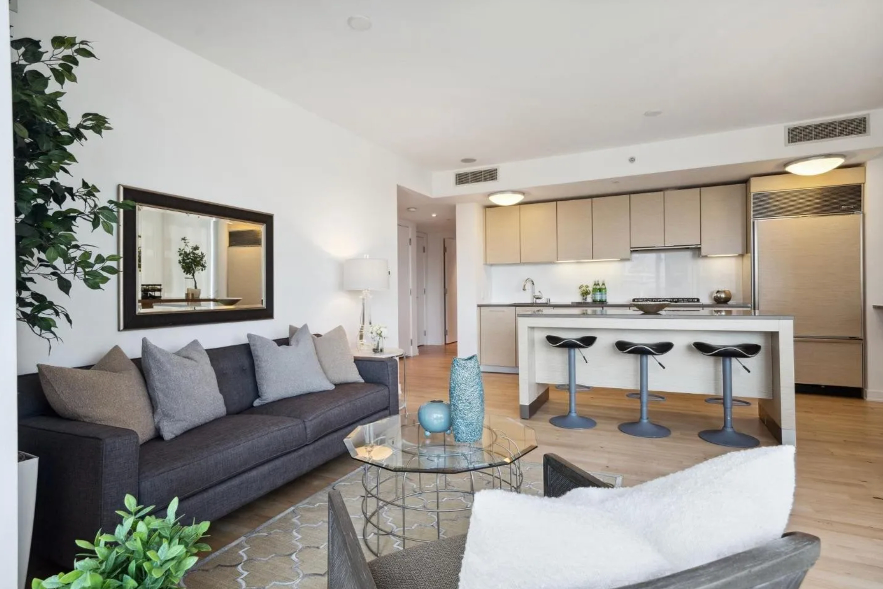 A modern living room and kitchen features a gray sofa with pillows, a glass coffee table, bar stools at a kitchen island, light cabinets, and a large mirror on the wall.