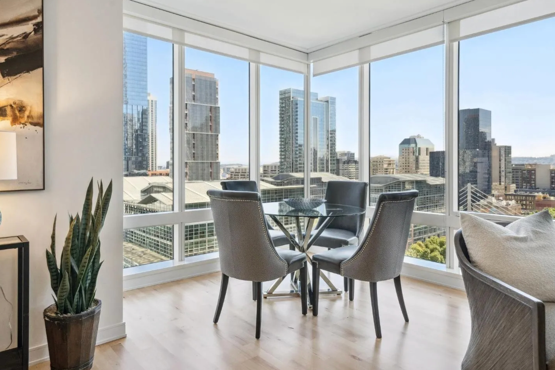 A modern dining area with a round glass table and four gray chairs is set in a corner of a high-rise apartment, offering a panoramic view of city skyscrapers.