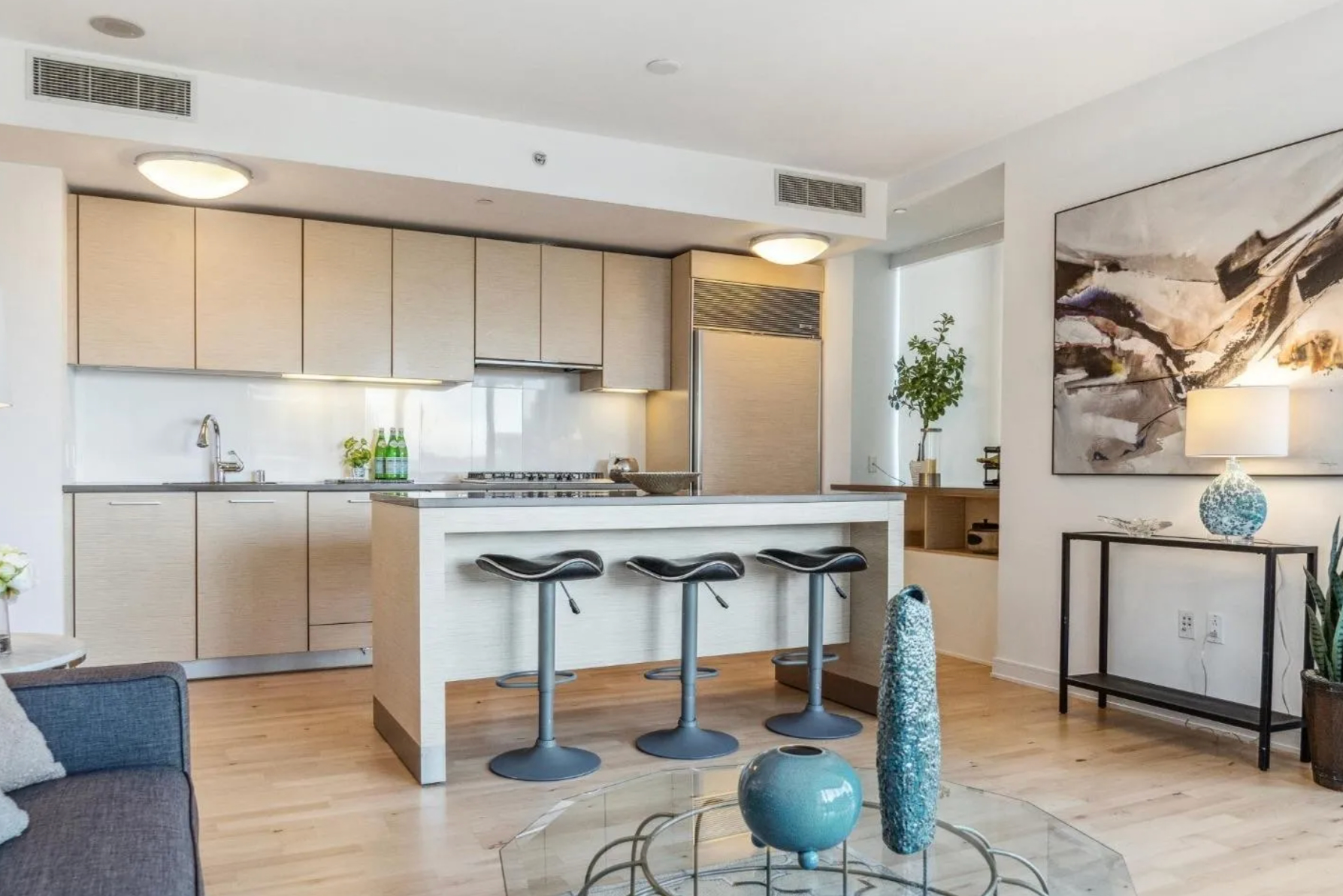 This image shows a modern kitchen with light wood cabinets, a white backsplash, and stainless steel appliances. There's an island with three barstools, and the kitchen opens into a cozy living area with abstract art and decorative items.