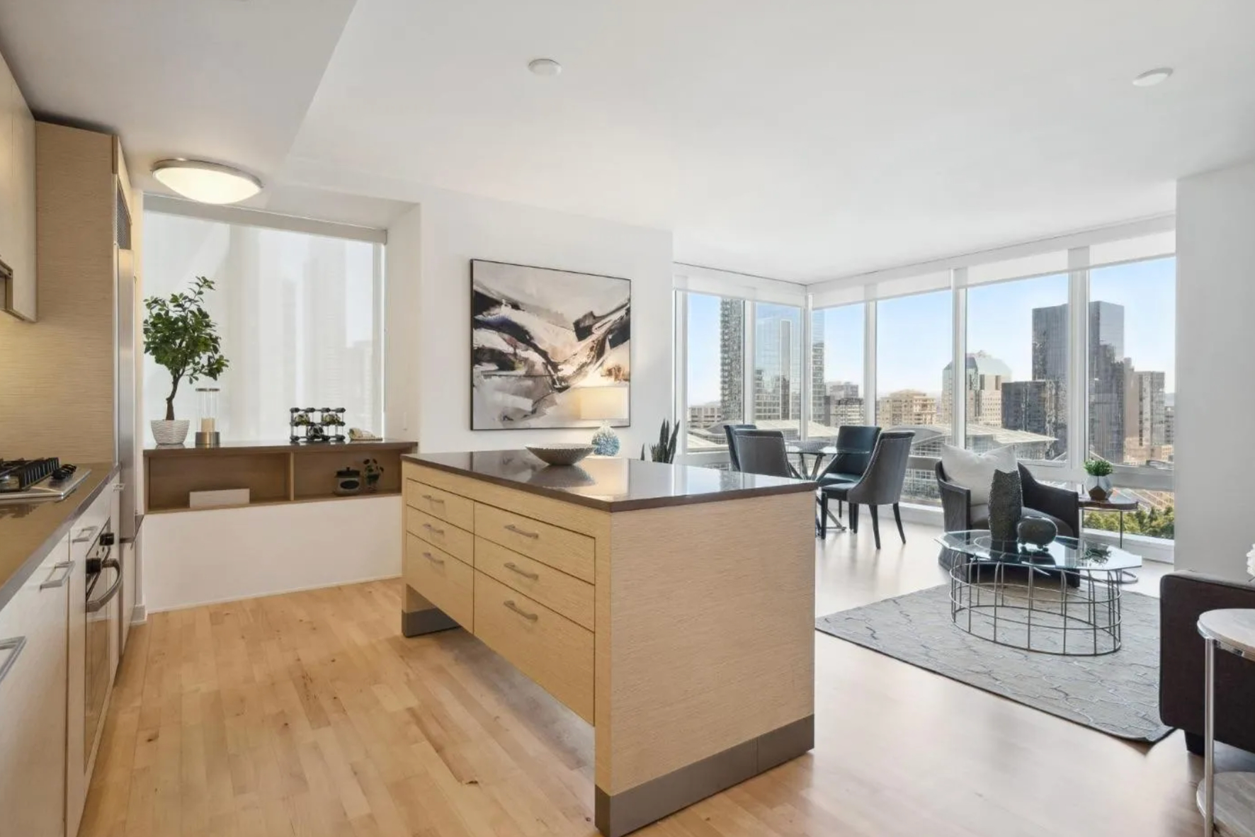 A modern kitchen and living area with a large island, light wood floors, abstract art, and floor-to-ceiling windows showcasing a city skyline.