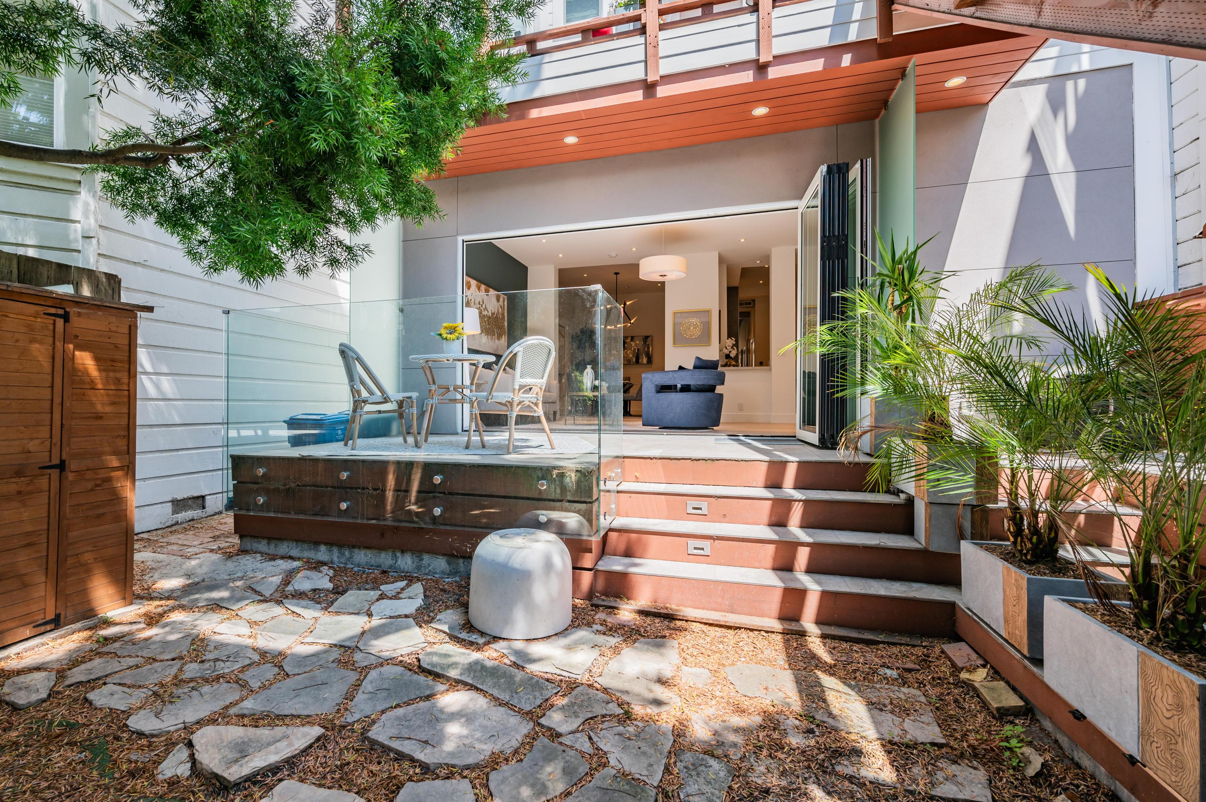 An open-concept living space with a small outdoor patio featuring a table and chairs. Steps lead to a modern, glass-enclosed living area with plants and a wooden storeroom.