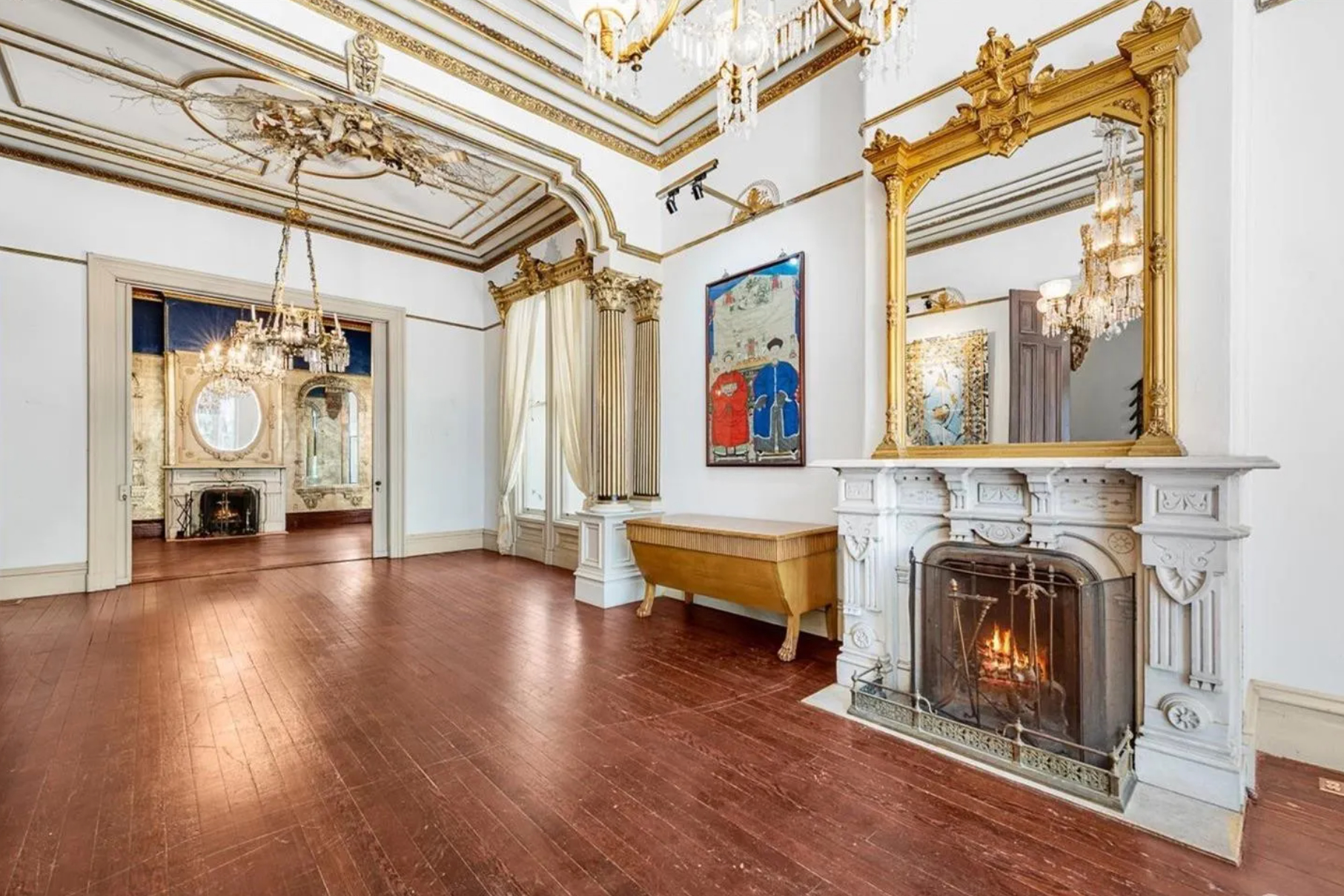 A grand room features an ornate fireplace with a large mirror, chandeliers, gold-trimmed white walls, wooden floors, and a glimpse of an adjoining space.