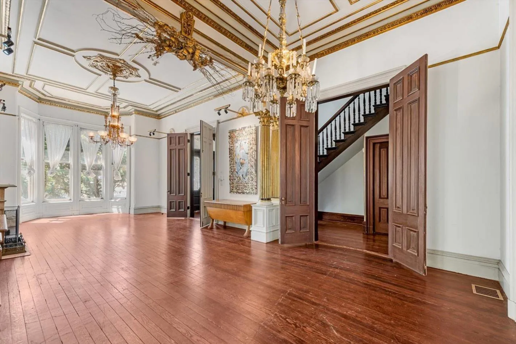 The image showcases an elegant, spacious room with polished hardwood floors, intricate moldings, chandeliers, large windows with sheer curtains, and a staircase in the background.