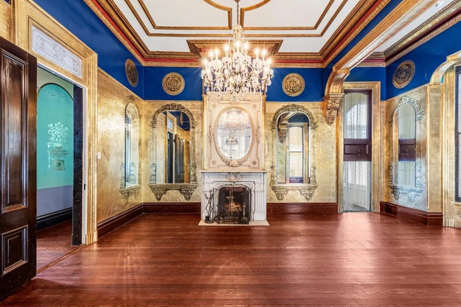 The ornate room features rich blue walls with gold accents, large mirrors, a crystal chandelier, a white fireplace, and hardwood floors which give a palatial feel.