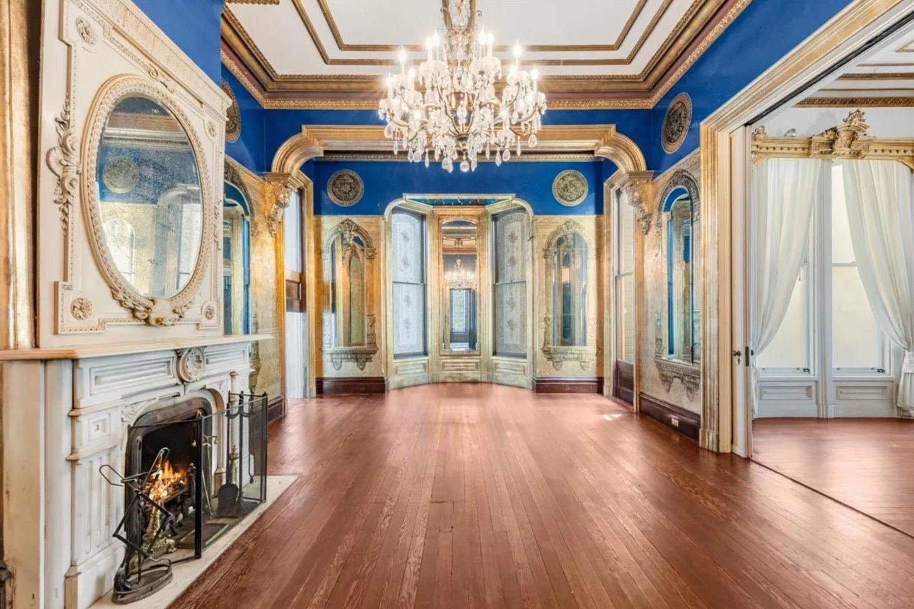 This room features elegant gold and blue walls, a wooden floor, chandeliers, arched windows, and a decorative white fireplace with a mirror above it.