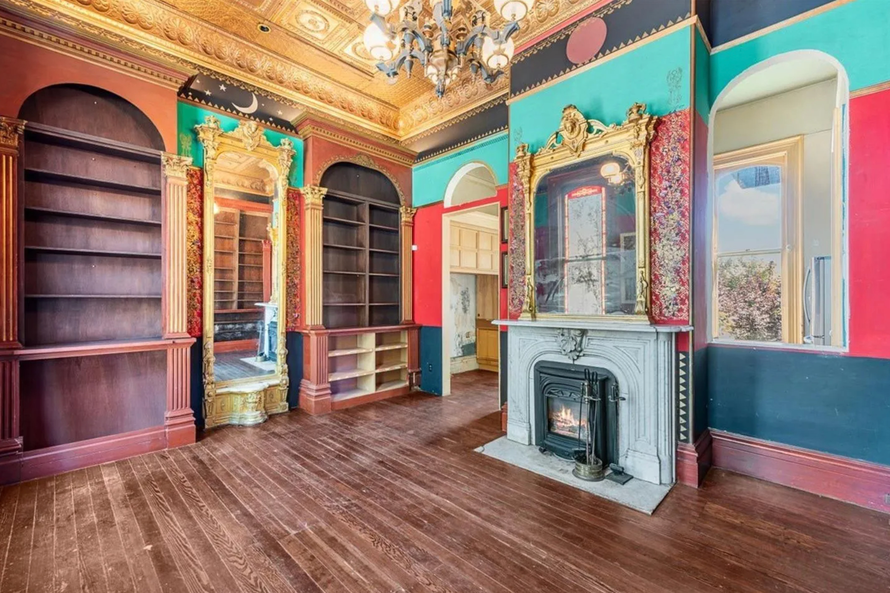 An ornate room features an elaborate ceiling, vibrant walls, empty wooden shelves, a decorative fireplace, a tall gold-framed mirror, and a chandelier on hardwood floors.