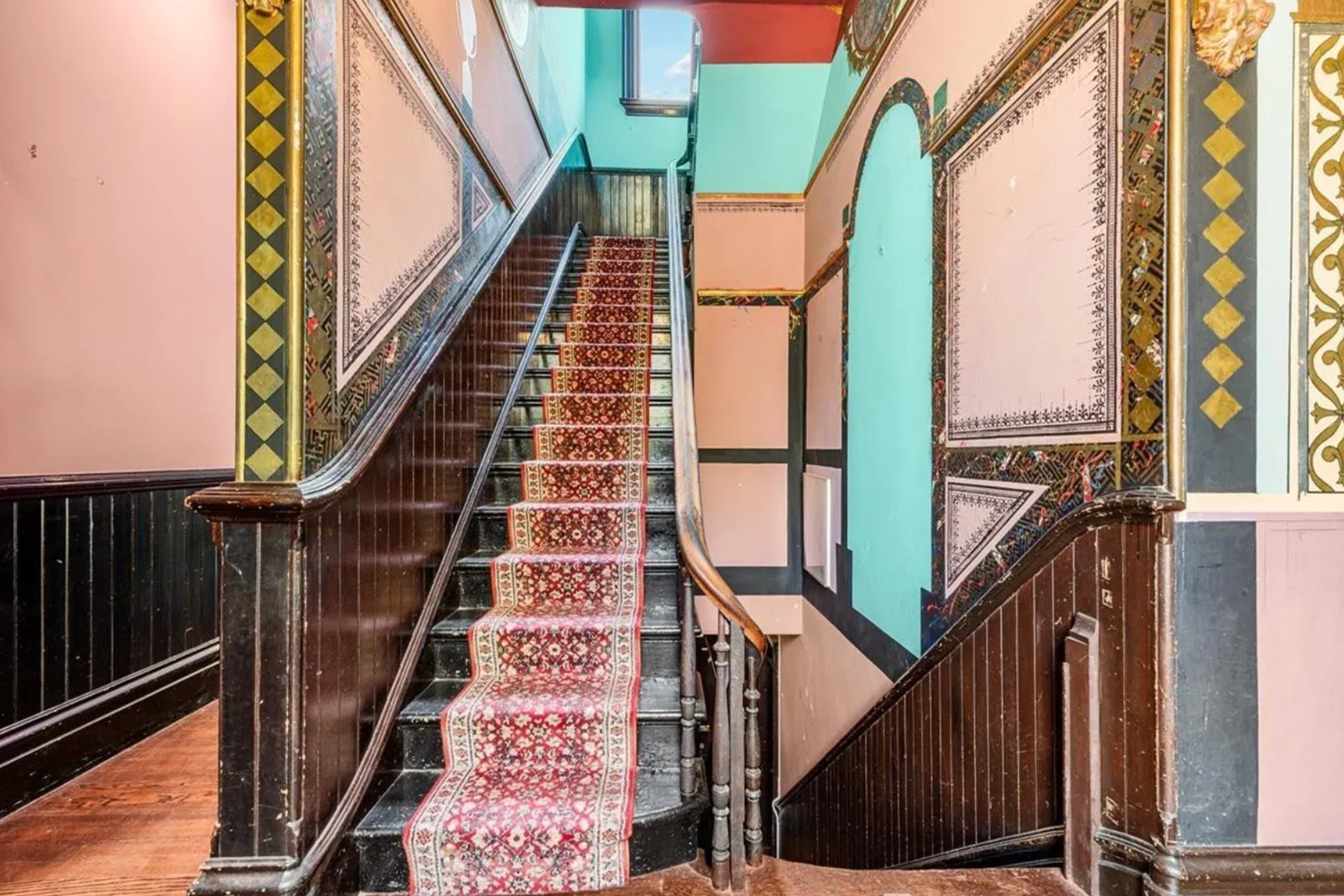 A staircase featuring a patterned red carpet leads upwards, flanked by ornate, colorful walls with intricate designs. A window at the top illuminates the area.