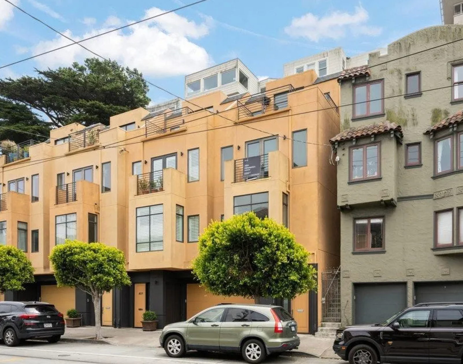 The image shows a row of urban townhouses with modern and stucco exteriors; cars are parked on the street in front, and there are a few small trees along the sidewalk.