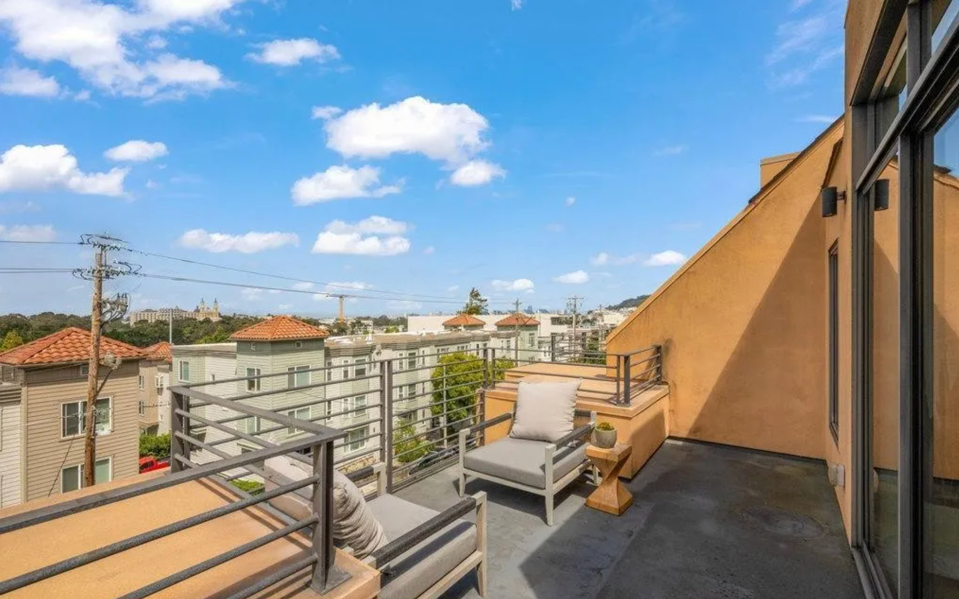 The image shows a rooftop patio with modern outdoor furniture, overlooking a cityscape of buildings with terracotta roofs, under a clear blue sky with scattered clouds.