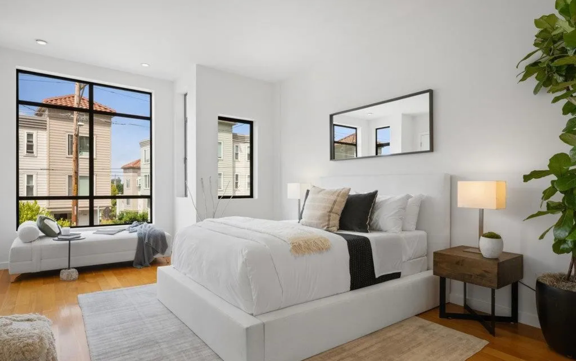 A bright, modern bedroom with large windows, a cozy bed, a bench by the window, a bedside table with a lamp, and a potted plant. Decor is minimalist in style.