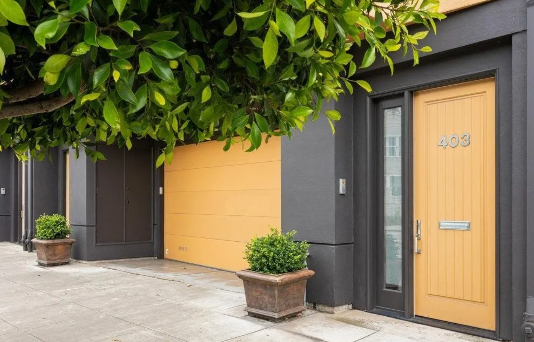 A grey building with yellow doors, labeled 403, has a large garage door and an entry door. Potted plants and a leafy tree overhang are also visible nearby.