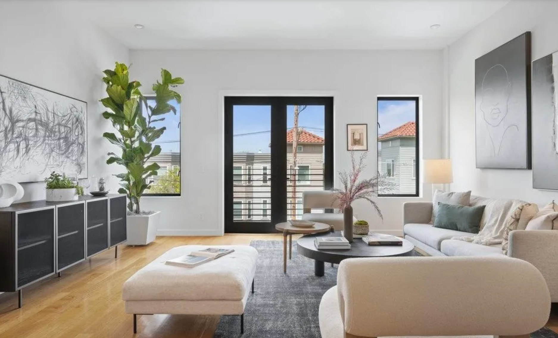 A bright living room with a beige sofa, modern art, and plants, includes a comfy ottoman, nested tables, and large windows letting in natural light.